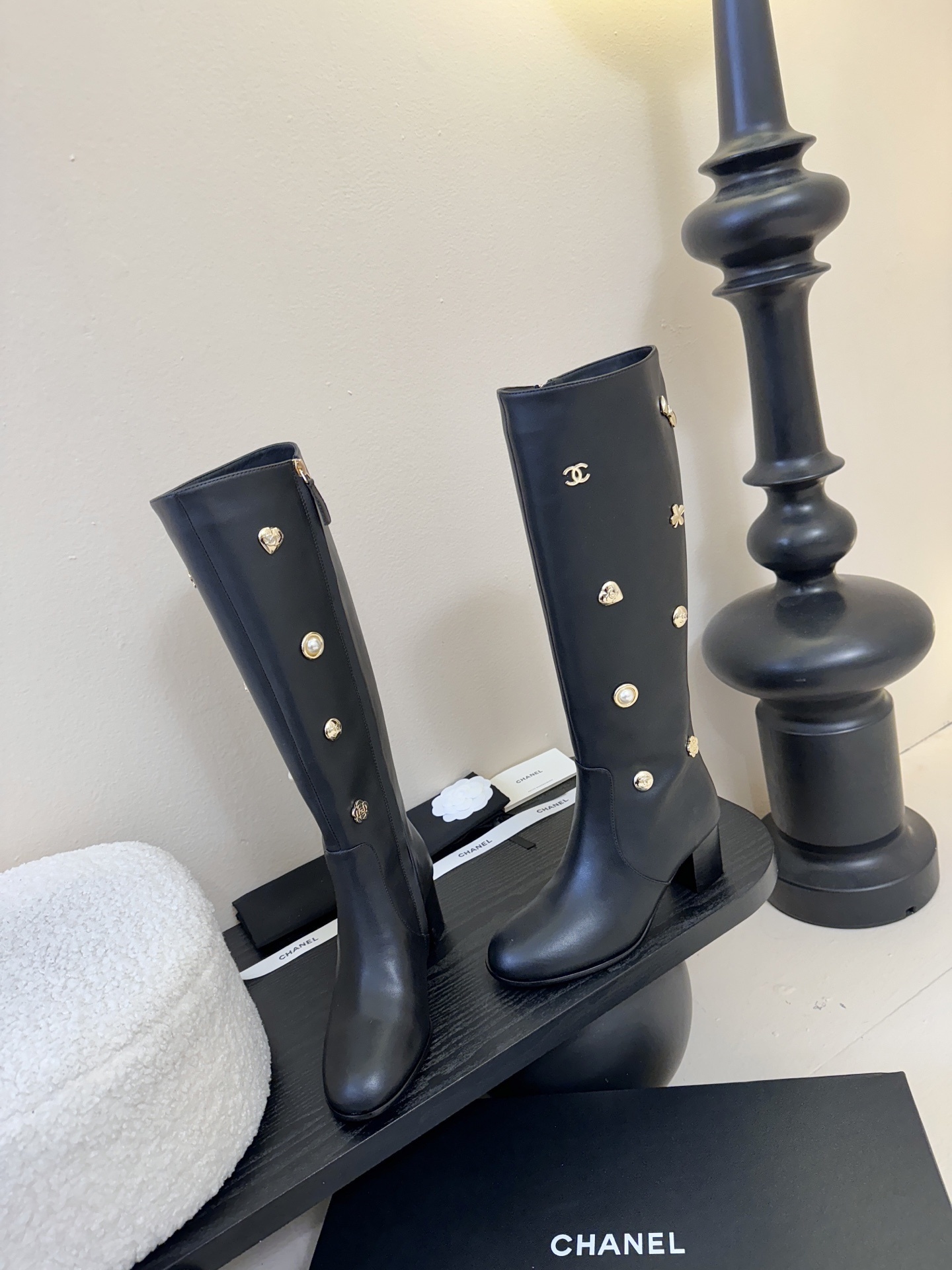 Pair of Chanel black leather knee-high boots decorated with gold CC logos, pearls, and heart charms on a black display. - Hình 3