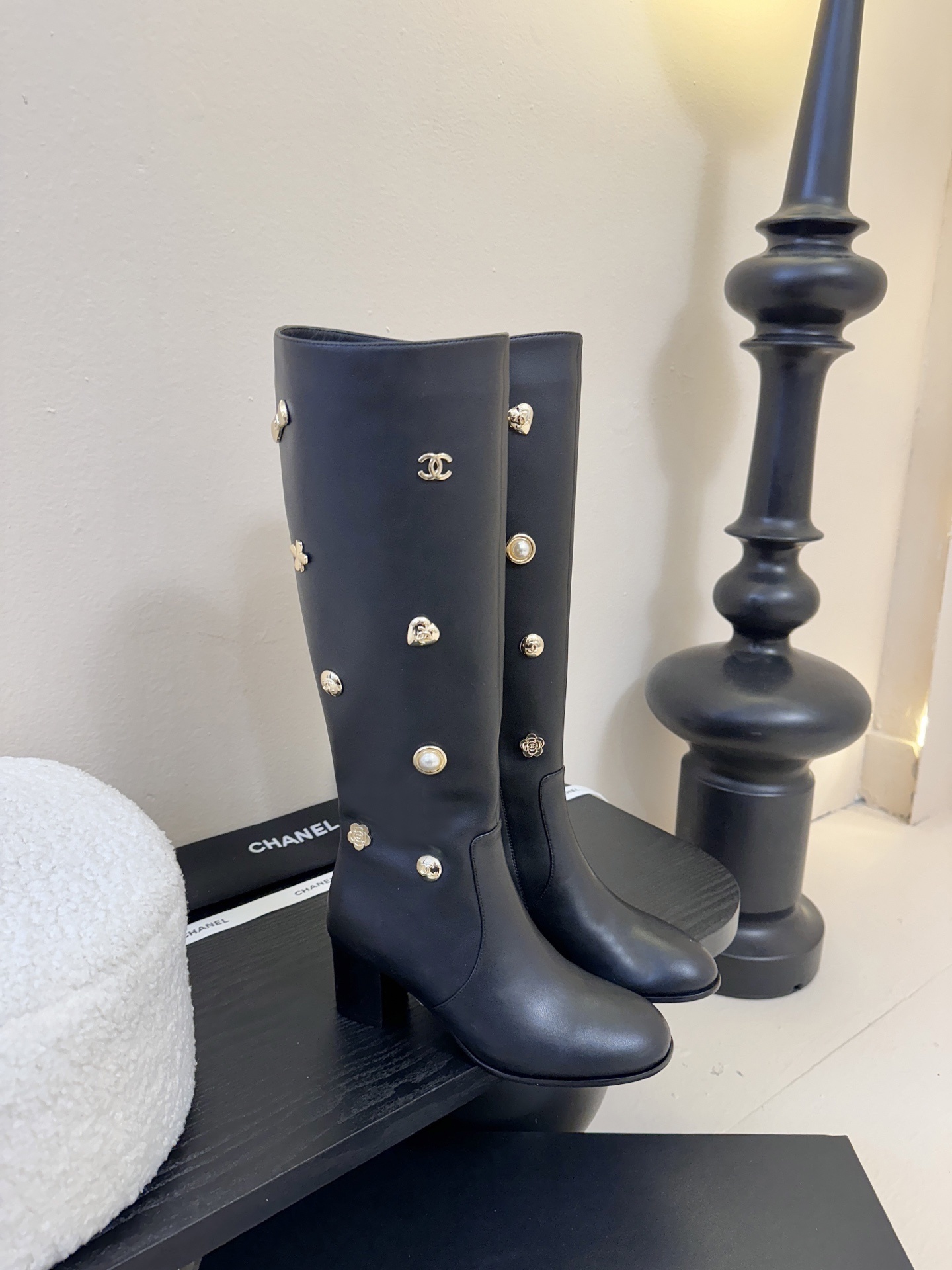 Pair of Chanel black leather knee-high boots decorated with gold CC logos, pearls, and heart charms on a black display. - Hình 2