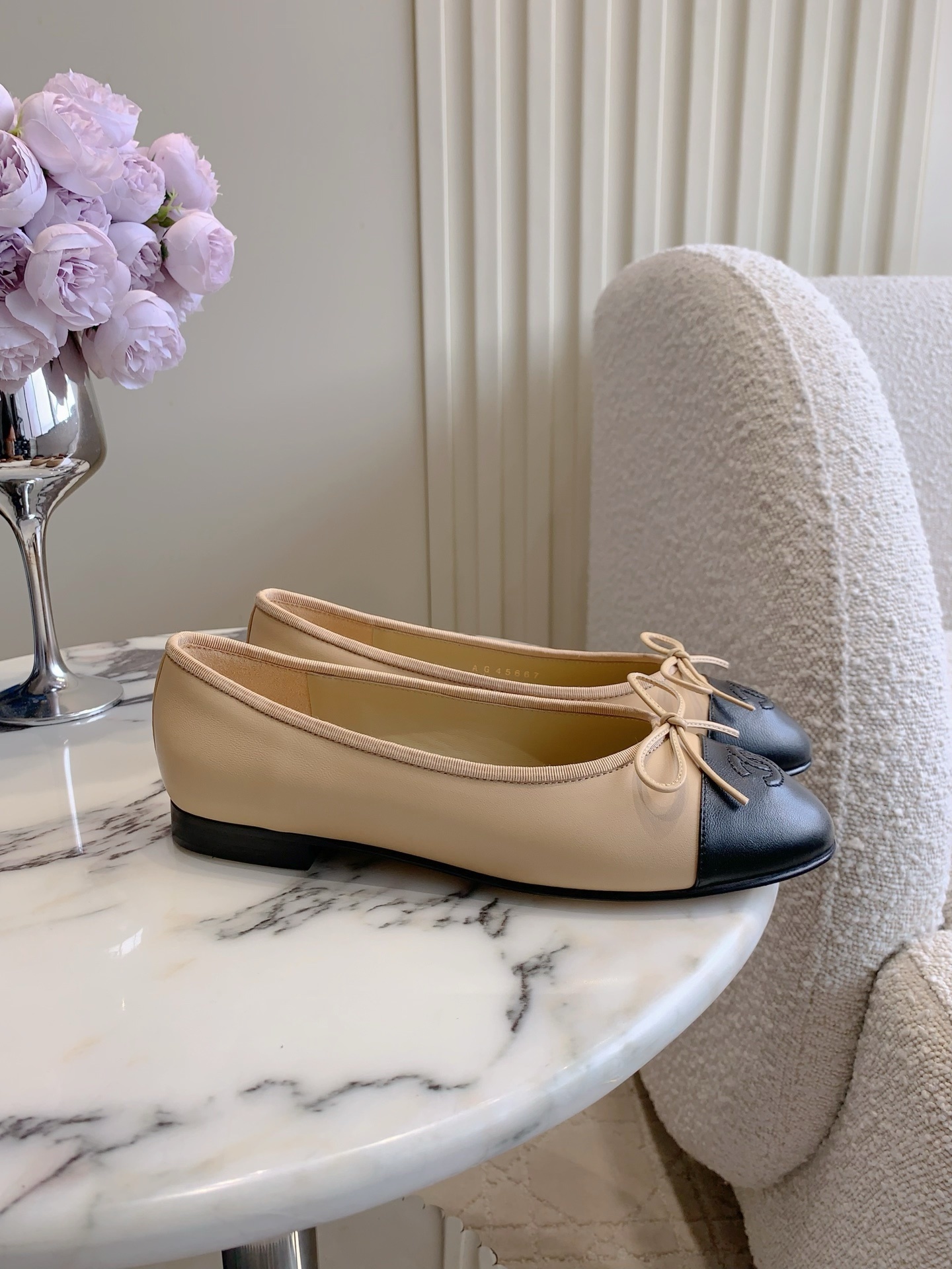 Classic Chanel beige lambskin ballerina flats with black toe caps and bow detail displayed on a marble surface. - Hình 2