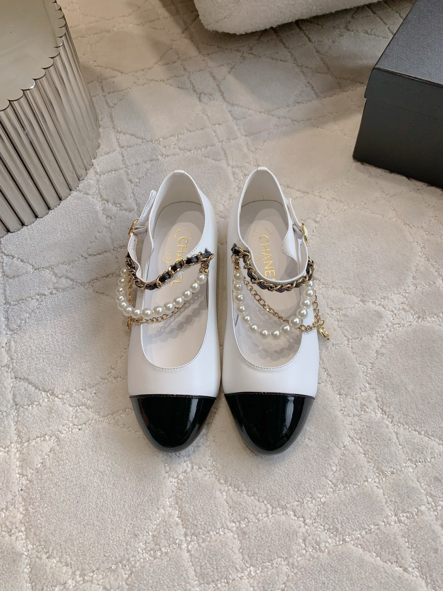Elegant white leather ballet flats with black patent cap toe and decorative pearl and gold chain strap on a marble surface - Hình 2