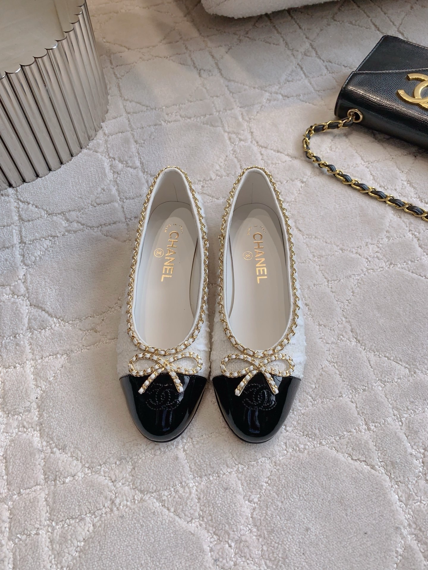 Pair of Chanel white tweed ballet flats with black patent leather cap toes and gold chain bow detailing on a white carpet - Hình 2