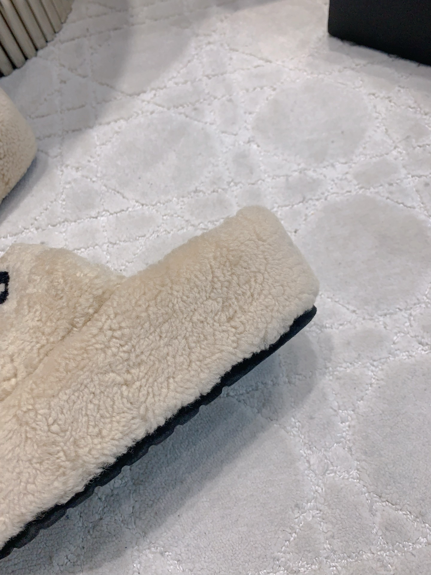 A pair of cream-colored fuzzy shearling platform slides featuring a black interlocking logo, displayed on a marble table and white carpet. - Hình 9