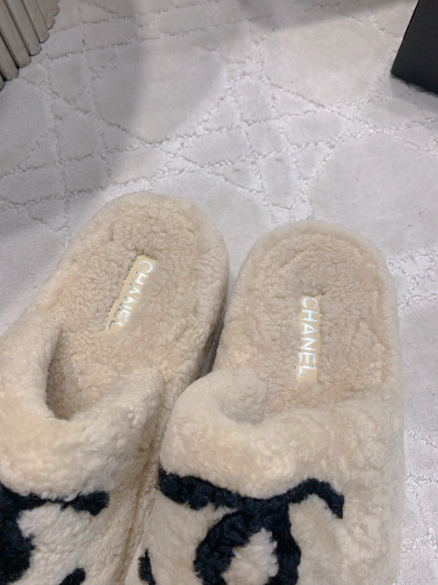 A pair of cream-colored fuzzy shearling platform slides featuring a black interlocking logo, displayed on a marble table and white carpet. - Hình 8