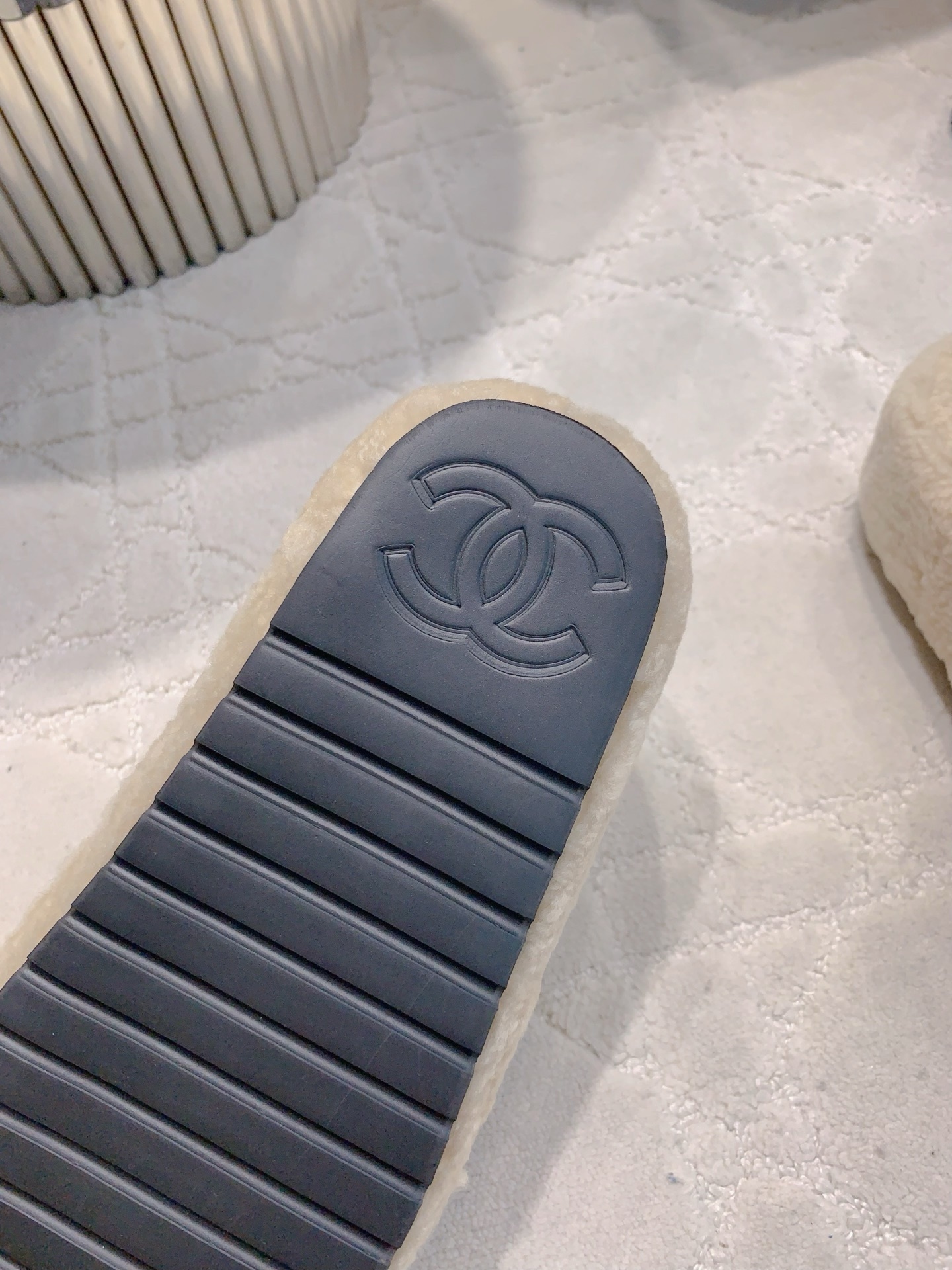 A pair of cream-colored fuzzy shearling platform slides featuring a black interlocking logo, displayed on a marble table and white carpet. - Hình 6