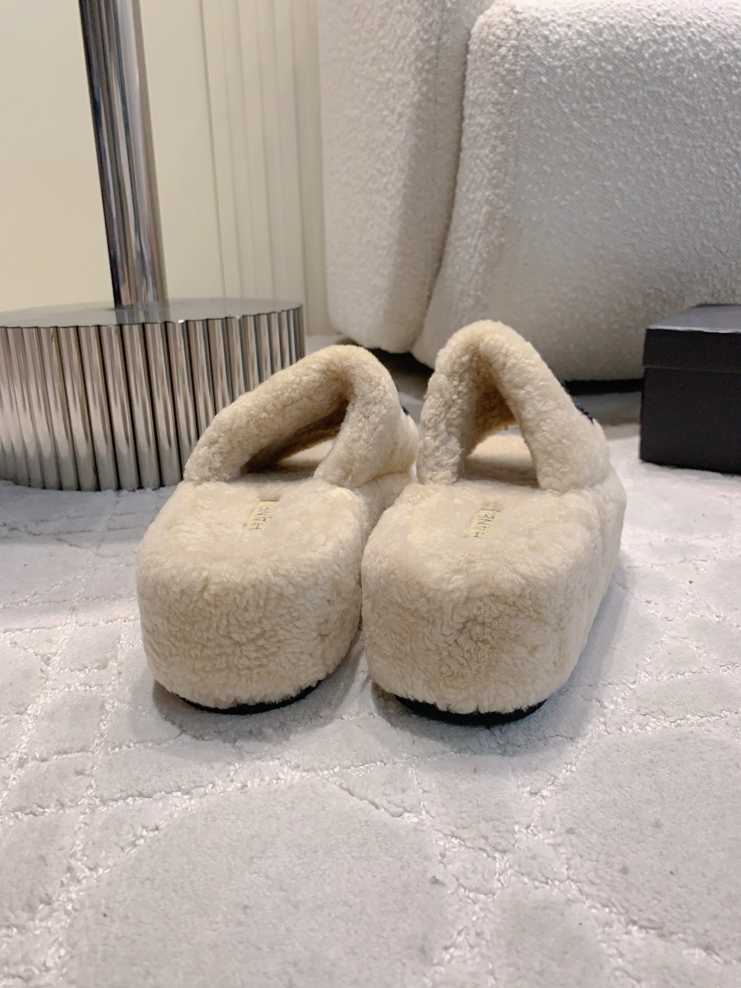 A pair of cream-colored fuzzy shearling platform slides featuring a black interlocking logo, displayed on a marble table and white carpet. - Hình 5