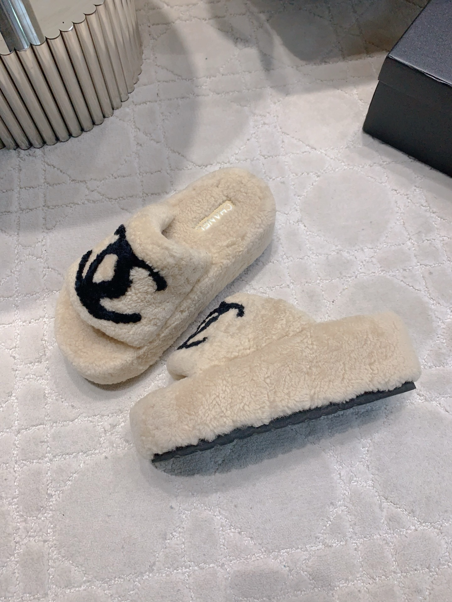 A pair of cream-colored fuzzy shearling platform slides featuring a black interlocking logo, displayed on a marble table and white carpet. - Hình 4