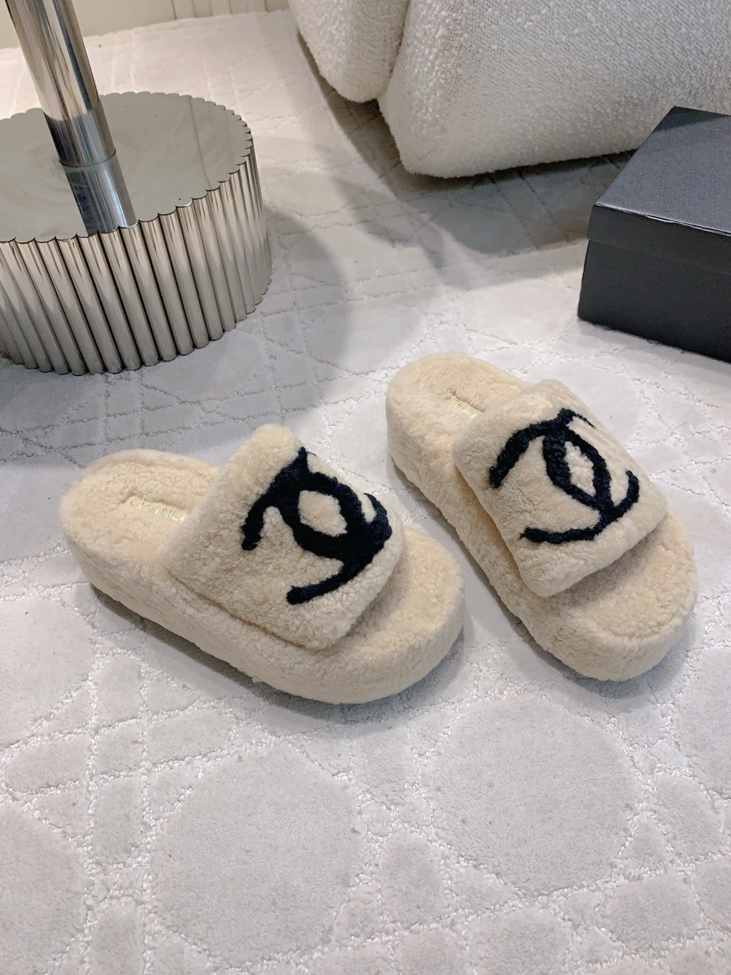 A pair of cream-colored fuzzy shearling platform slides featuring a black interlocking logo, displayed on a marble table and white carpet. - Hình 3