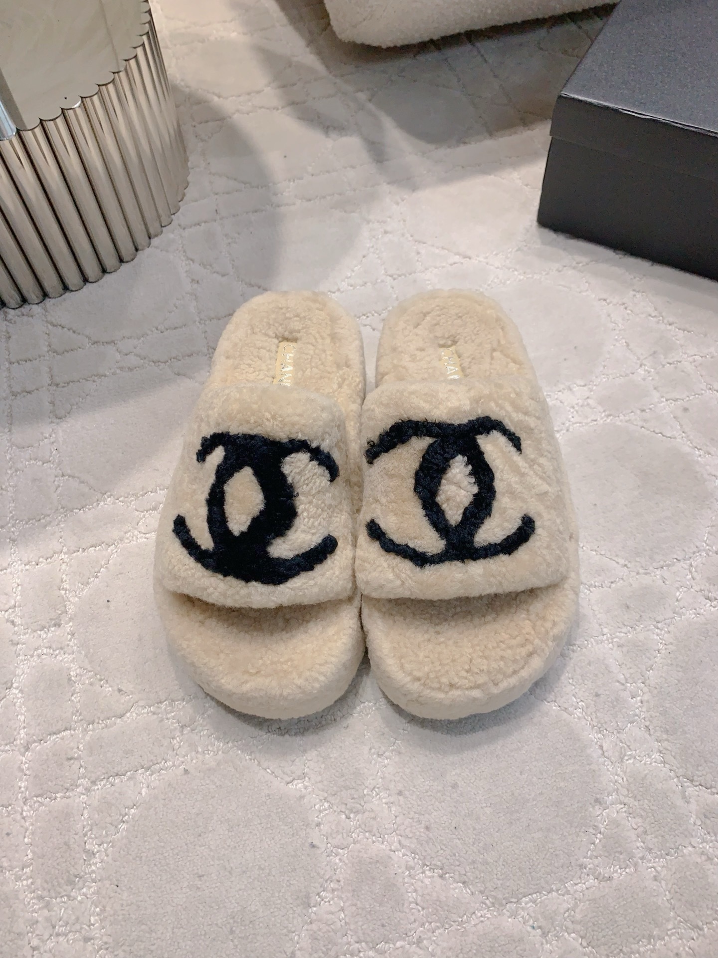 A pair of cream-colored fuzzy shearling platform slides featuring a black interlocking logo, displayed on a marble table and white carpet. - Hình 2