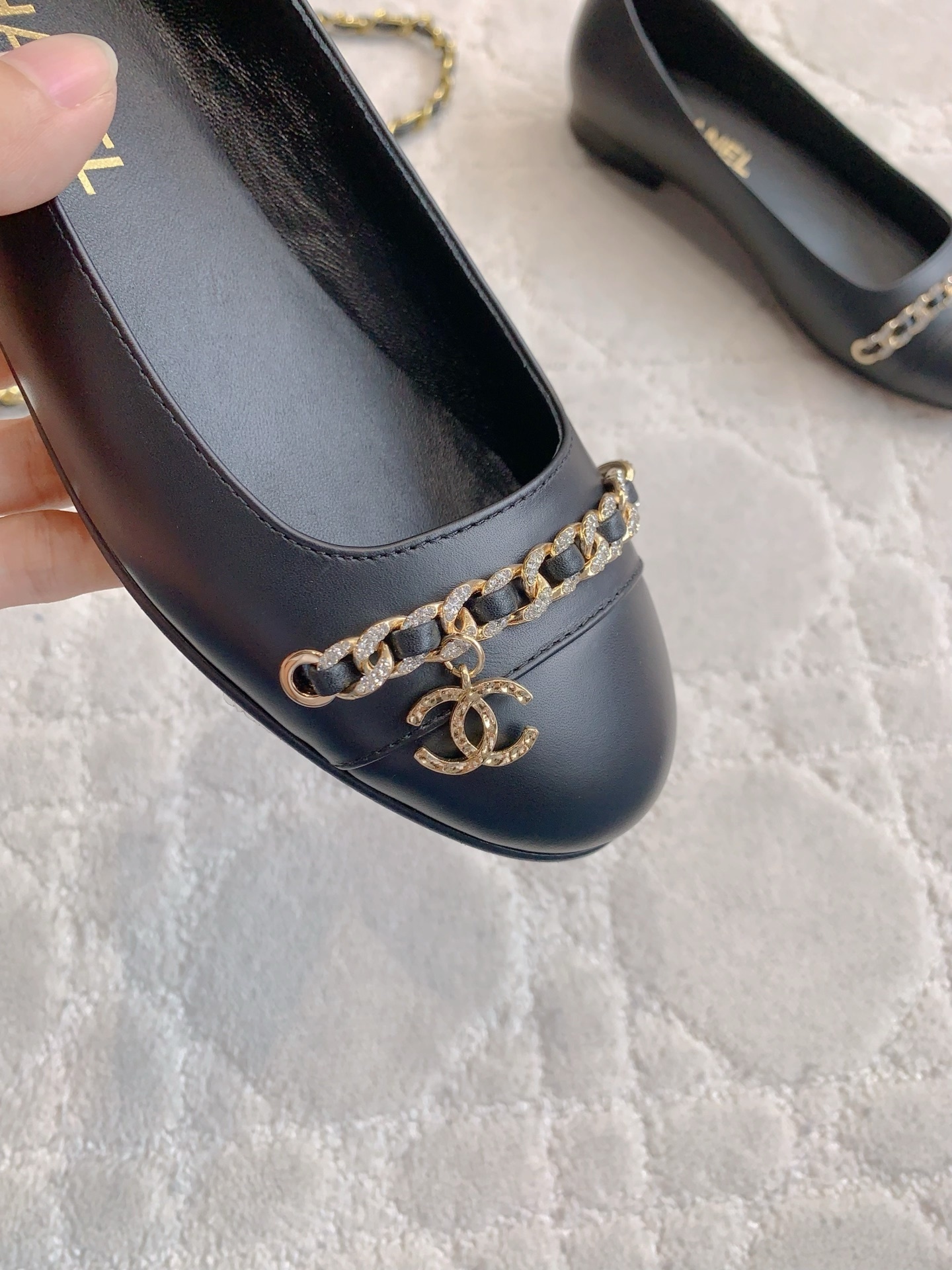 A pair of Chanel black leather ballerina flats with gold chain and CC logo charm displayed on a marble table. - Hình 7