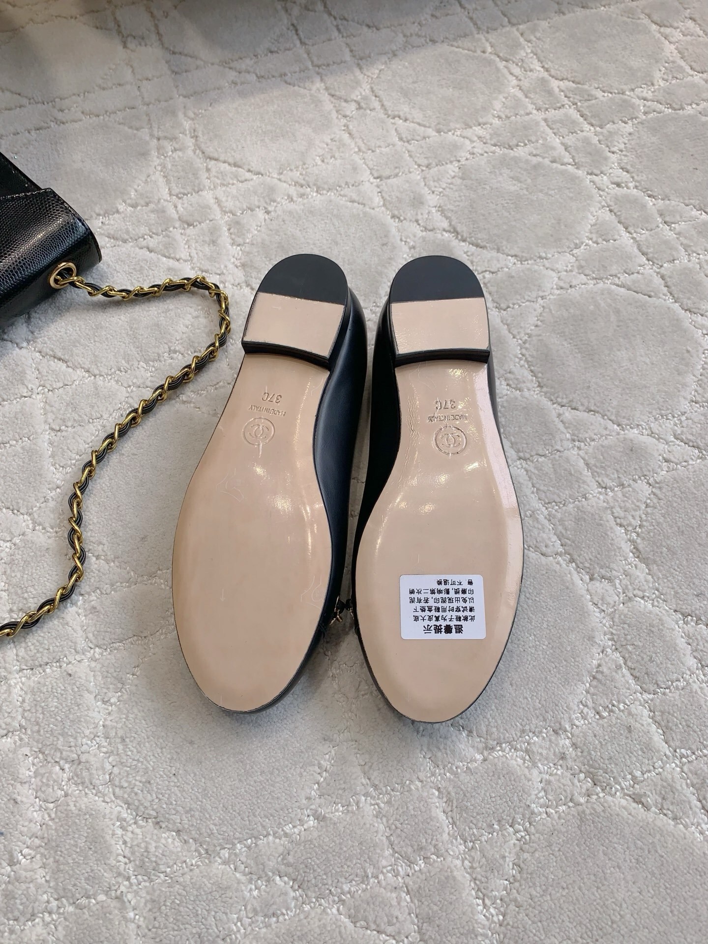 A pair of Chanel black leather ballerina flats with gold chain and CC logo charm displayed on a marble table. - Hình 6