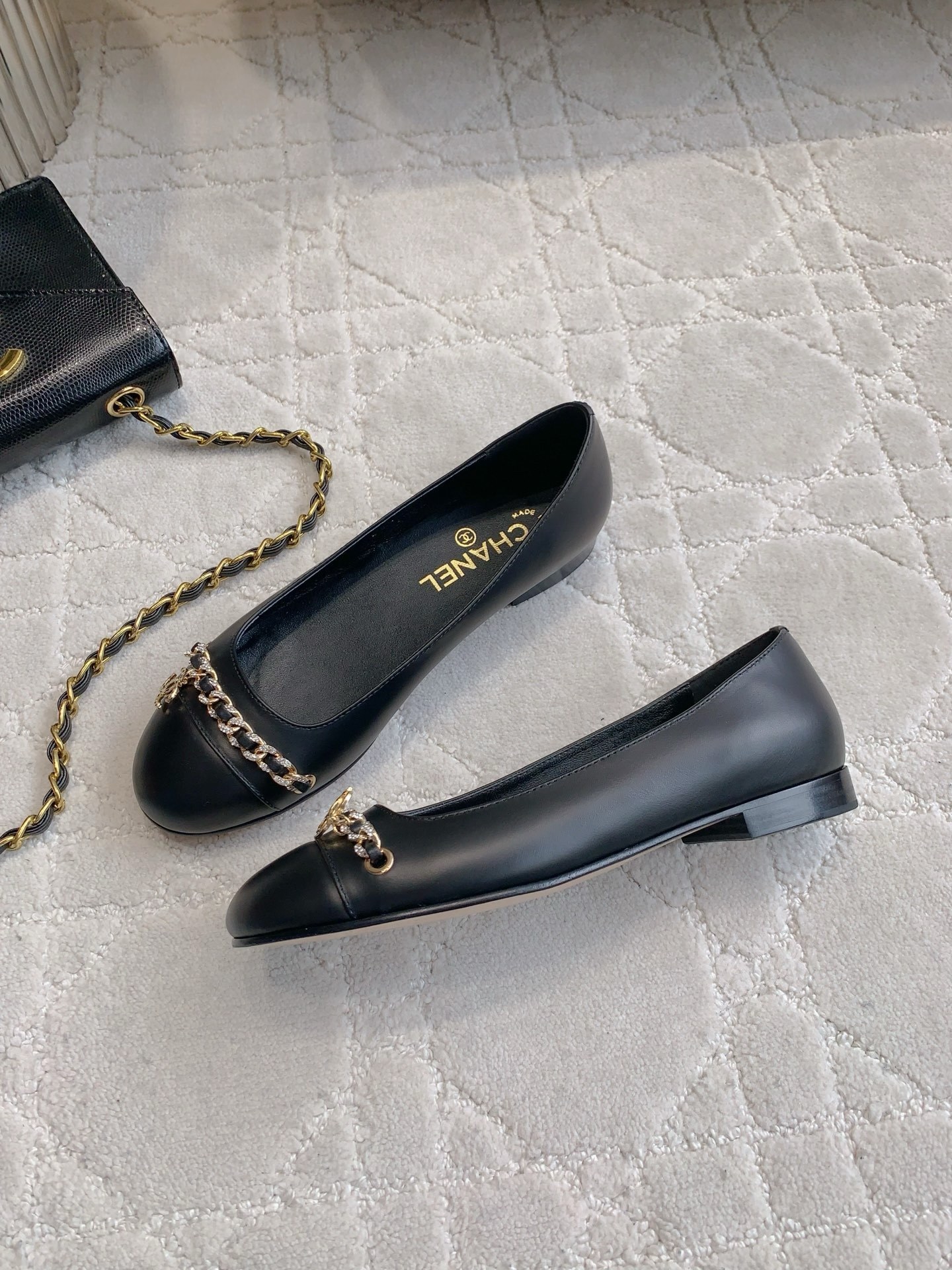 A pair of Chanel black leather ballerina flats with gold chain and CC logo charm displayed on a marble table. - Hình 4