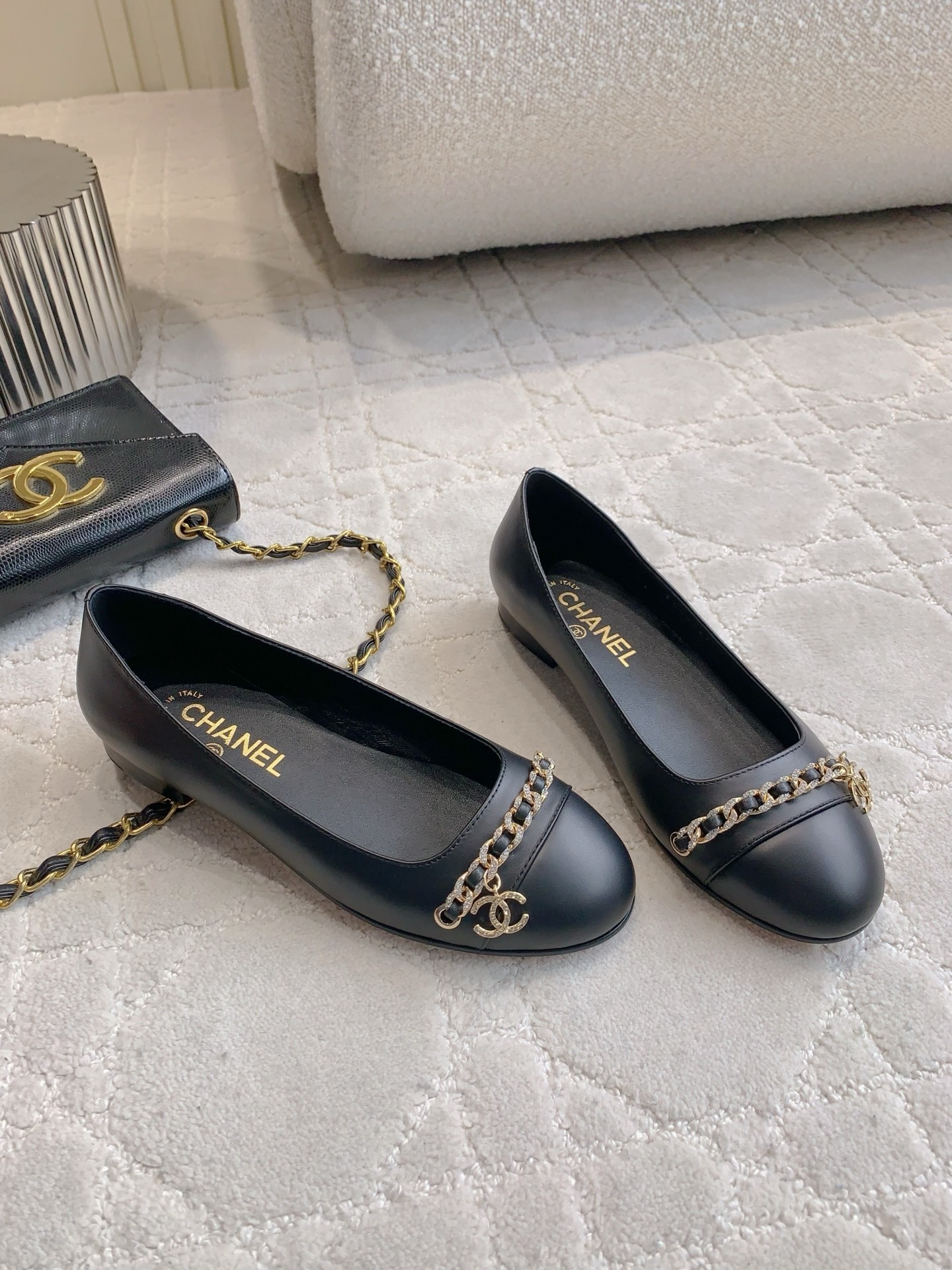 A pair of Chanel black leather ballerina flats with gold chain and CC logo charm displayed on a marble table. - Hình 3