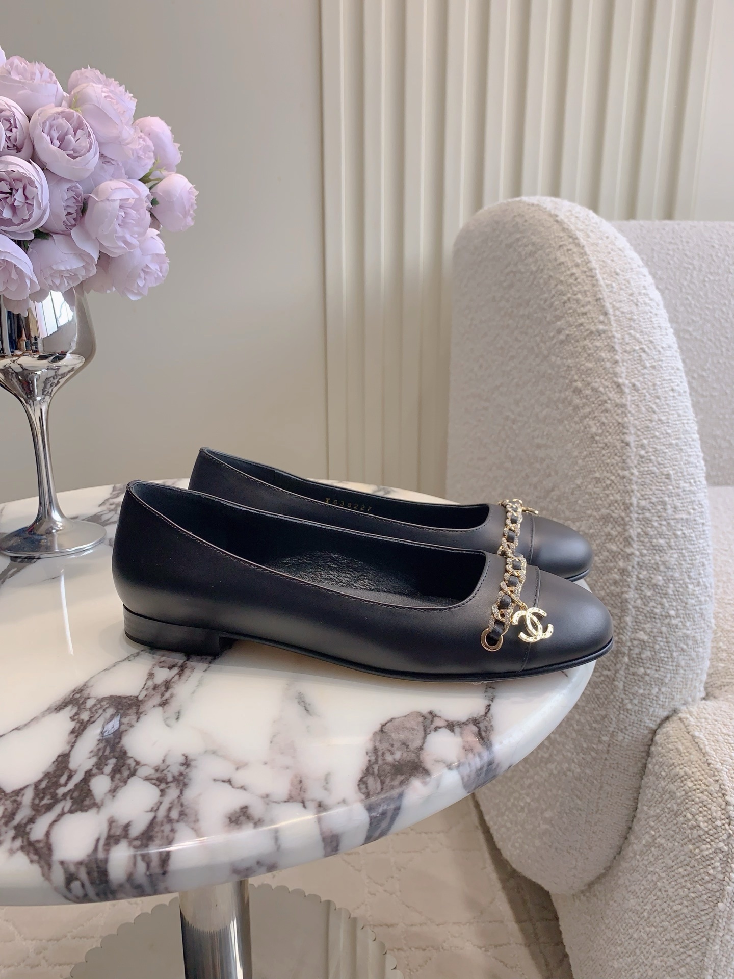 A pair of Chanel black leather ballerina flats with gold chain and CC logo charm displayed on a marble table. - Hình 2