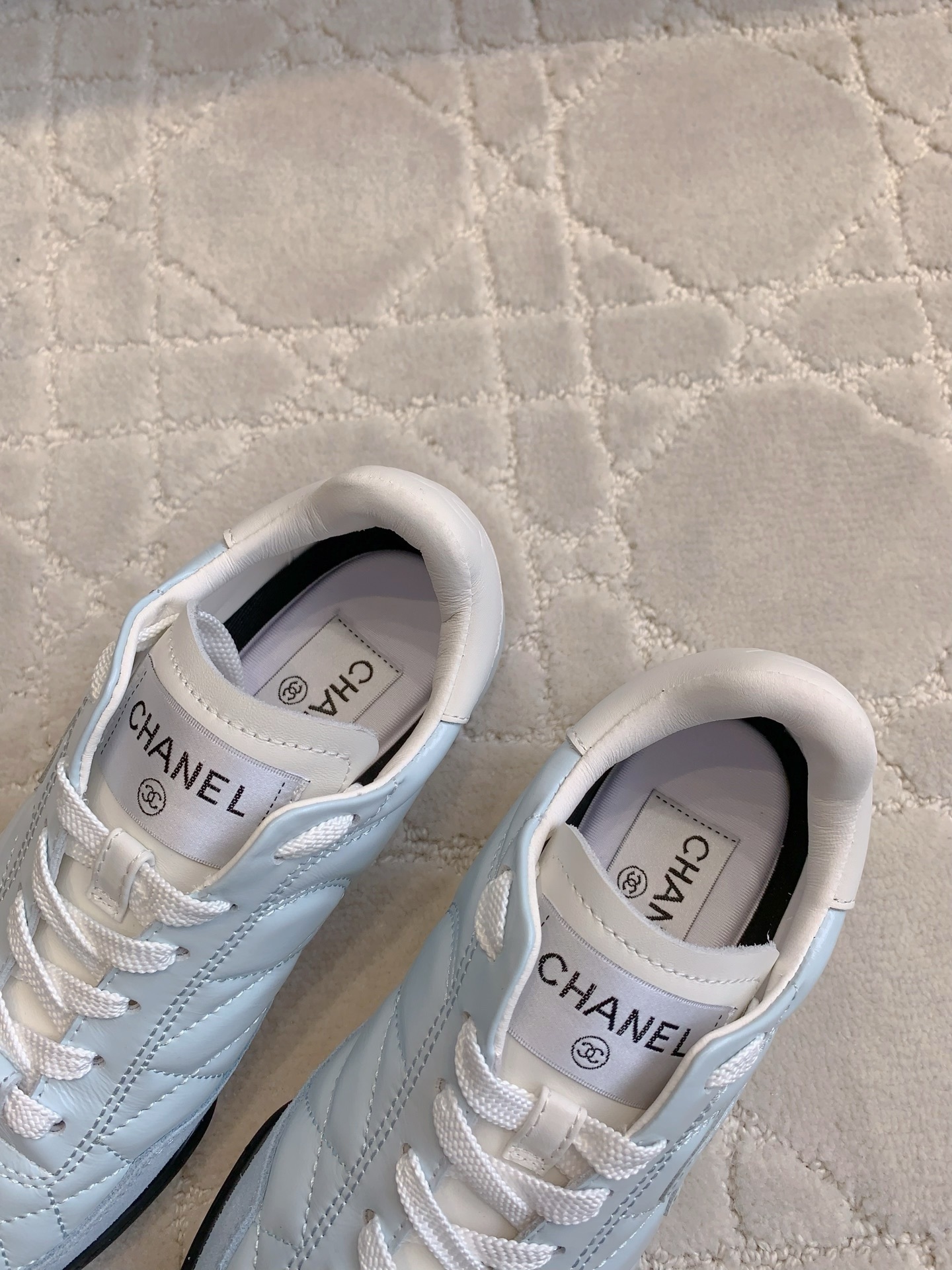 A pair of light blue Chanel quilted leather sneakers with suede toe caps and white laces displayed on a patterned carpet. - Hình 6