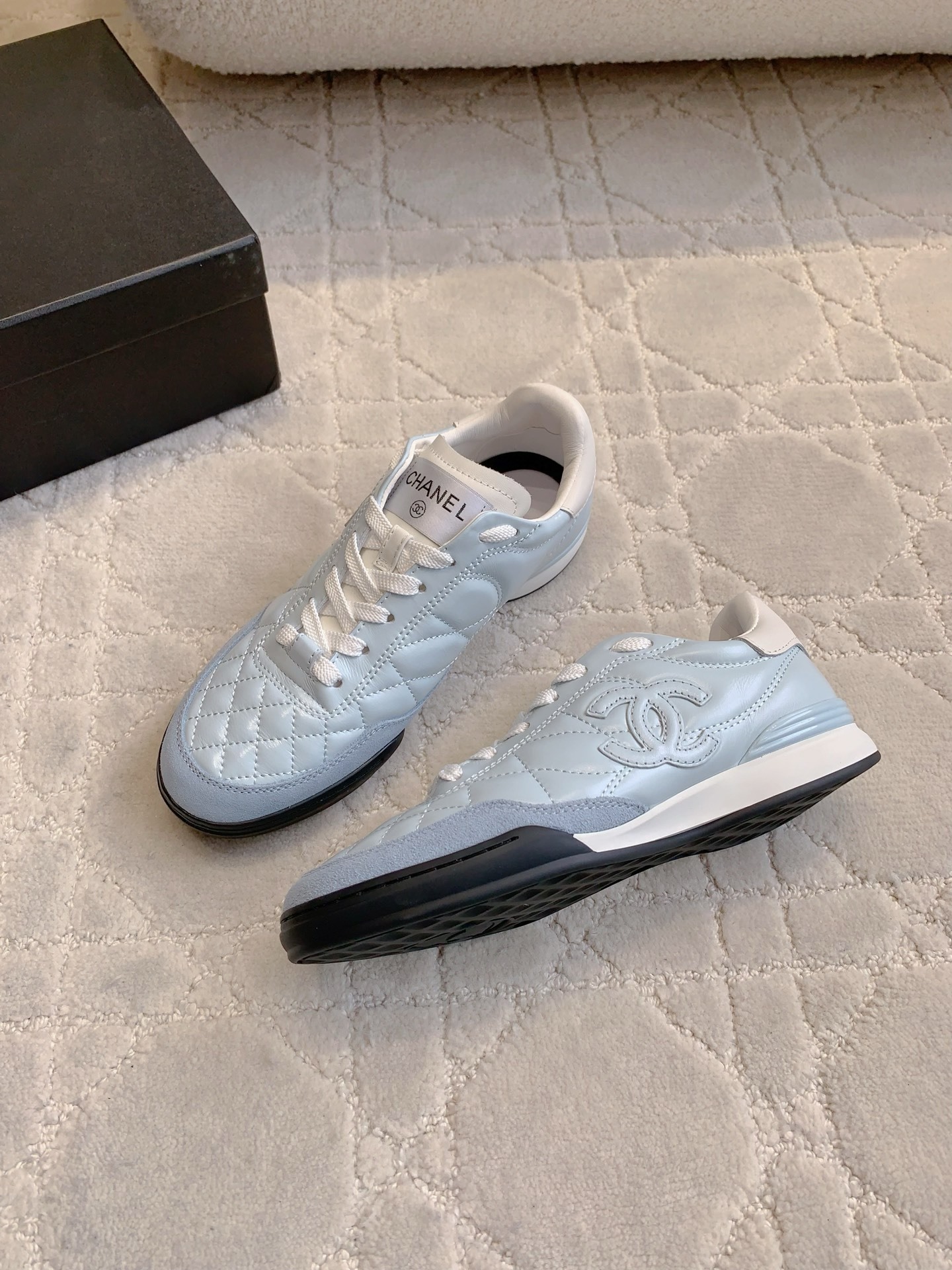 A pair of light blue Chanel quilted leather sneakers with suede toe caps and white laces displayed on a patterned carpet. - Hình 4