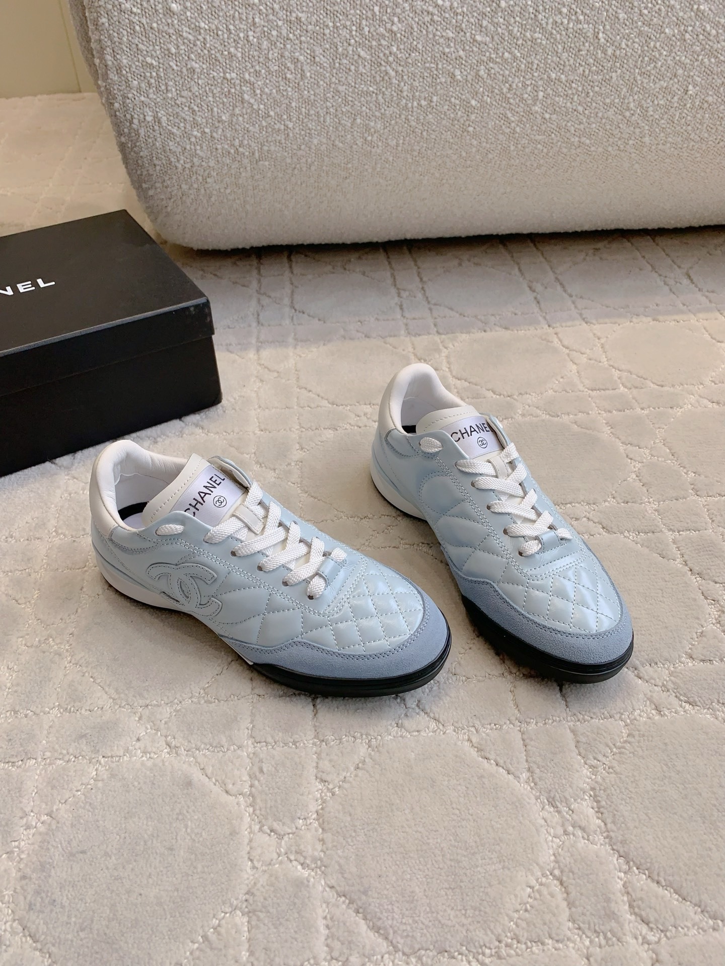 A pair of light blue Chanel quilted leather sneakers with suede toe caps and white laces displayed on a patterned carpet. - Hình 3