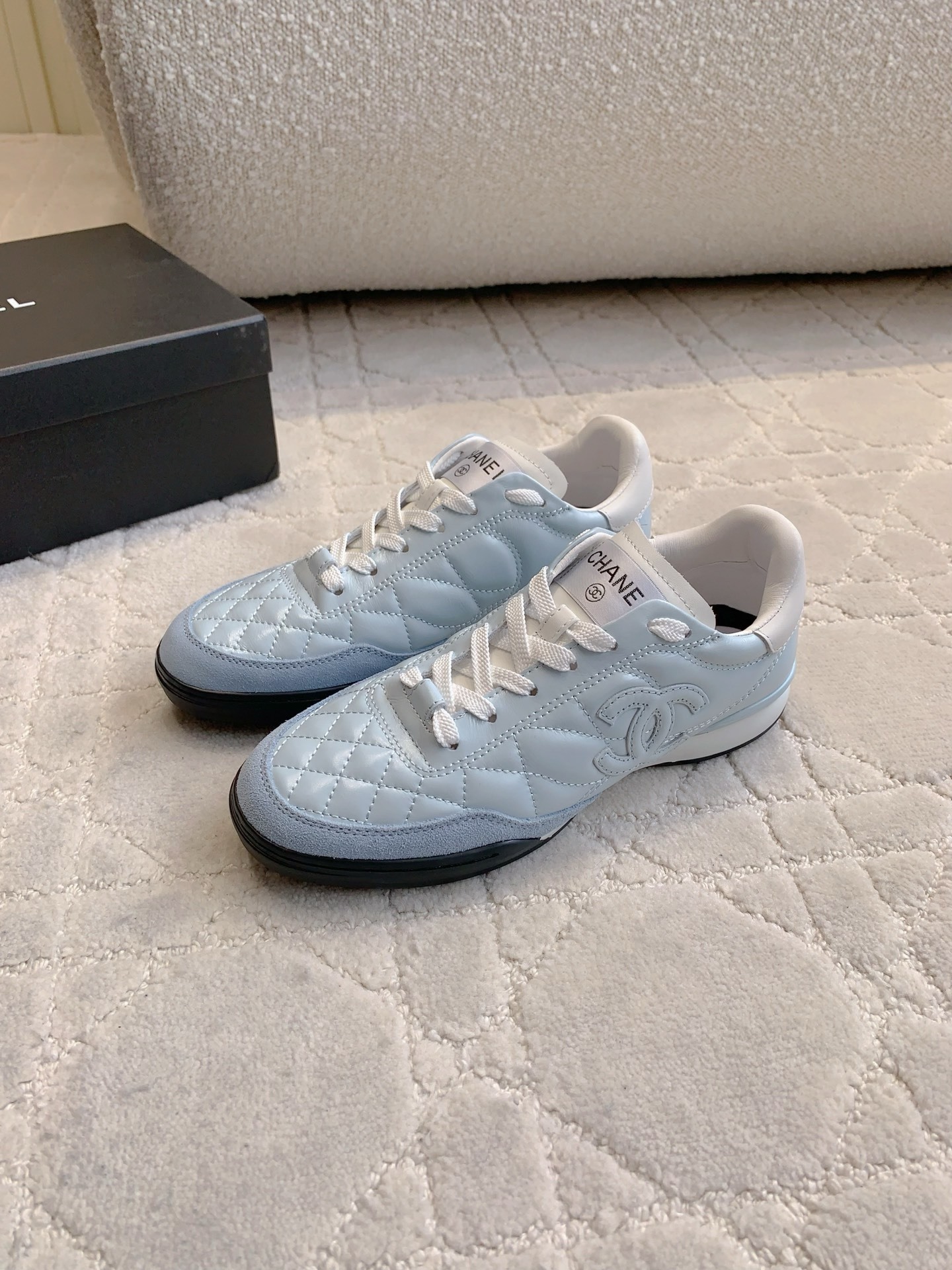 A pair of light blue Chanel quilted leather sneakers with suede toe caps and white laces displayed on a patterned carpet. - Hình 2