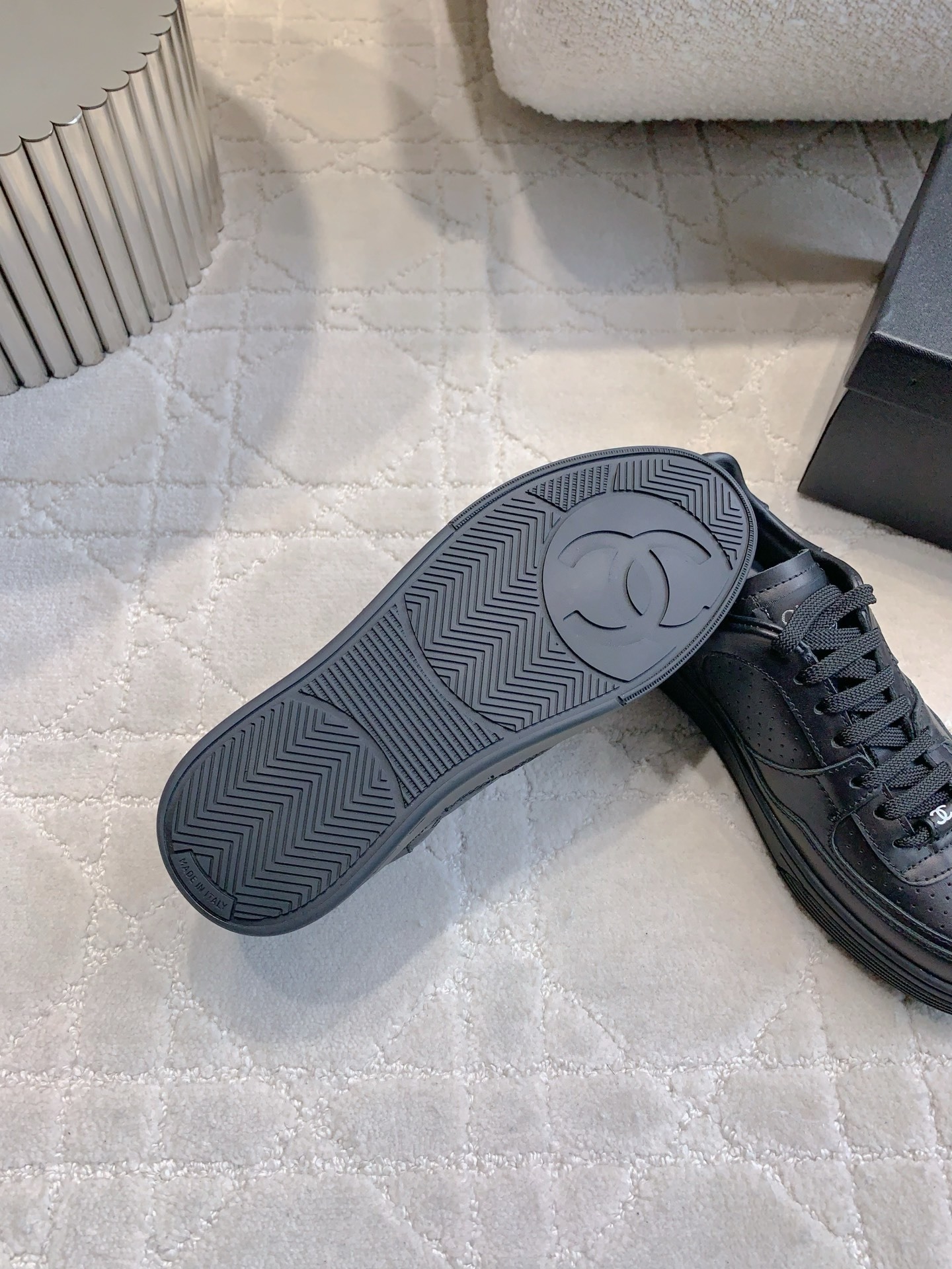 A pair of black Chanel leather sneakers featuring the interlocking CC logo and perforated details on a white textured carpet. - Hình 9