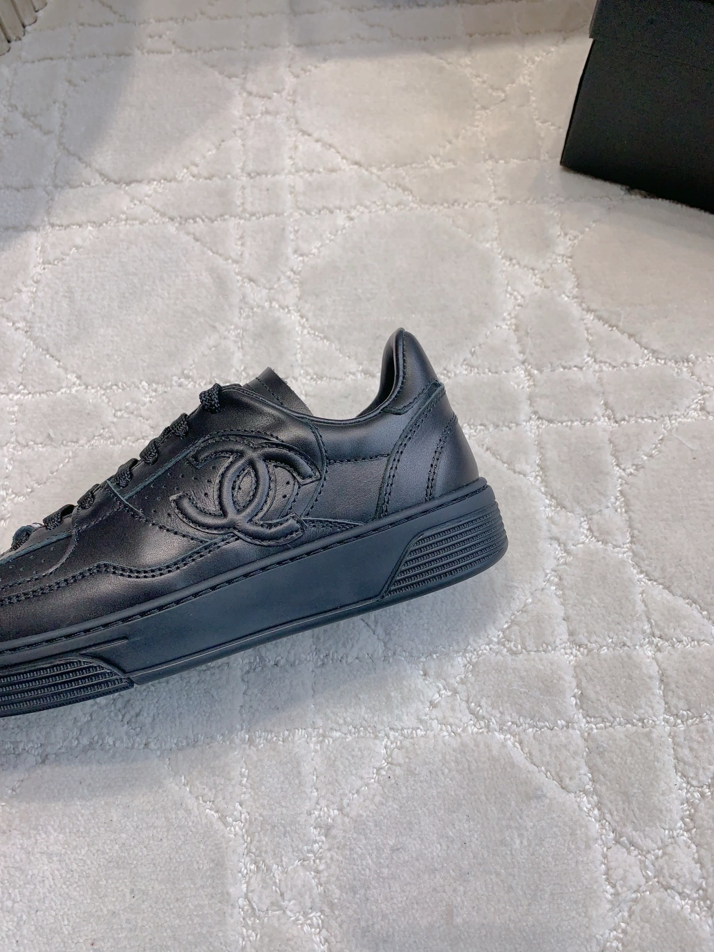 A pair of black Chanel leather sneakers featuring the interlocking CC logo and perforated details on a white textured carpet. - Hình 7