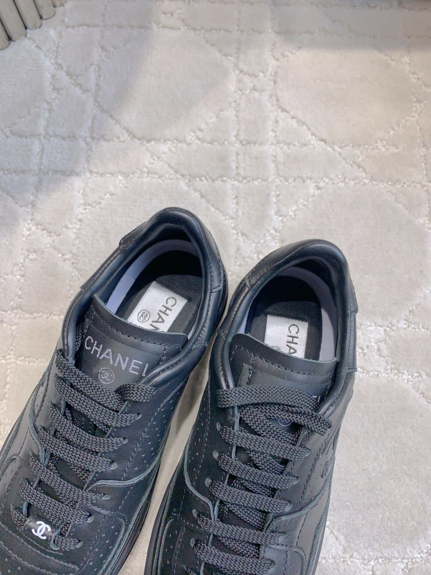 A pair of black Chanel leather sneakers featuring the interlocking CC logo and perforated details on a white textured carpet. - Hình 6