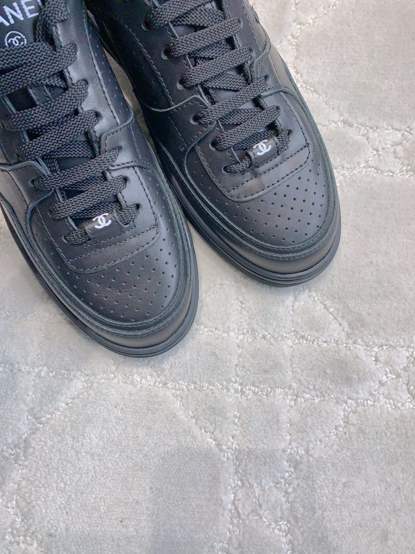 A pair of black Chanel leather sneakers featuring the interlocking CC logo and perforated details on a white textured carpet. - Hình 5