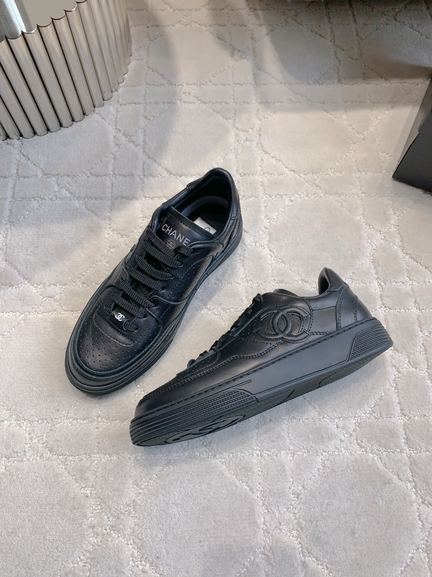 A pair of black Chanel leather sneakers featuring the interlocking CC logo and perforated details on a white textured carpet. - Hình 4