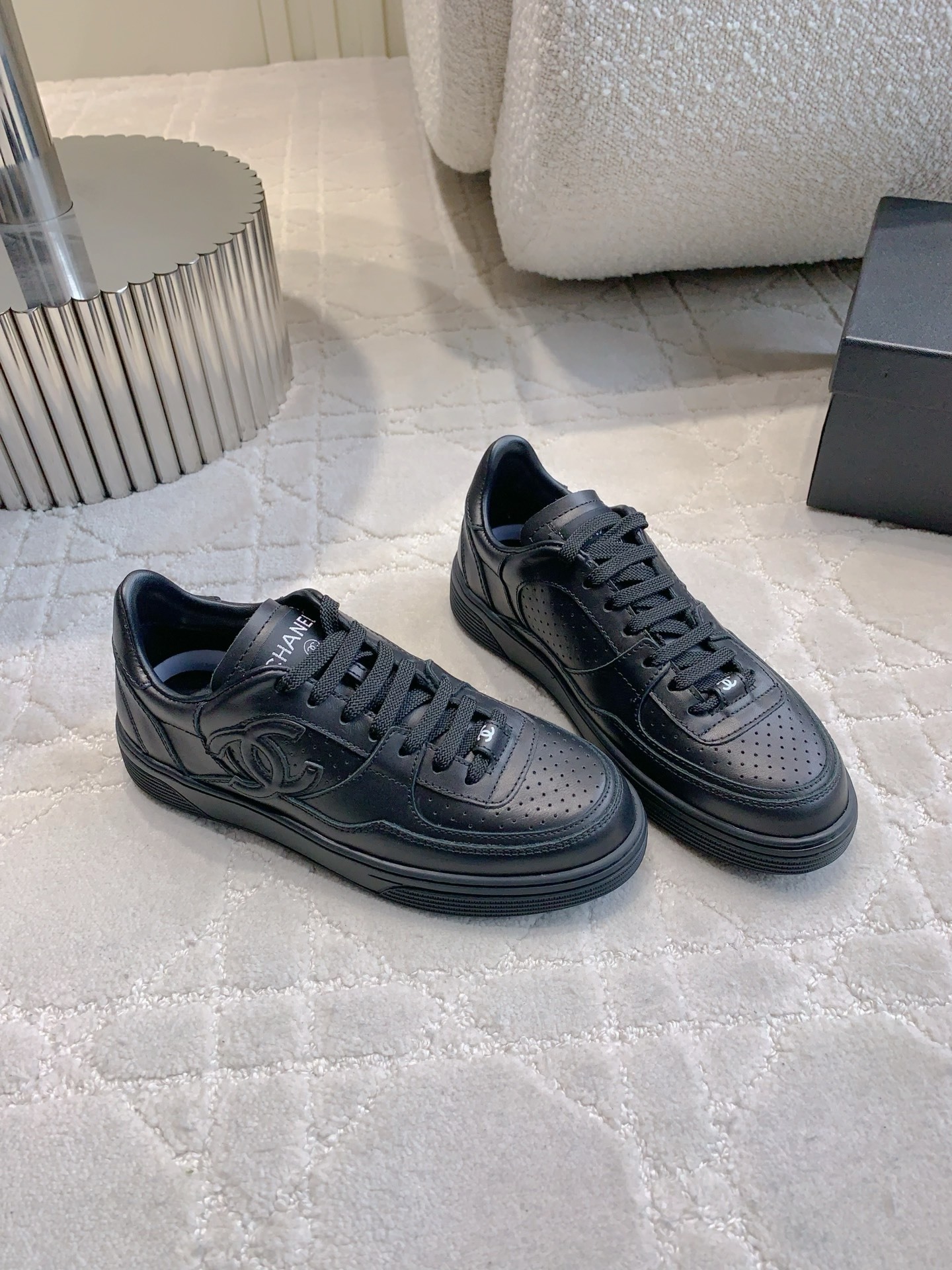 A pair of black Chanel leather sneakers featuring the interlocking CC logo and perforated details on a white textured carpet. - Hình 3