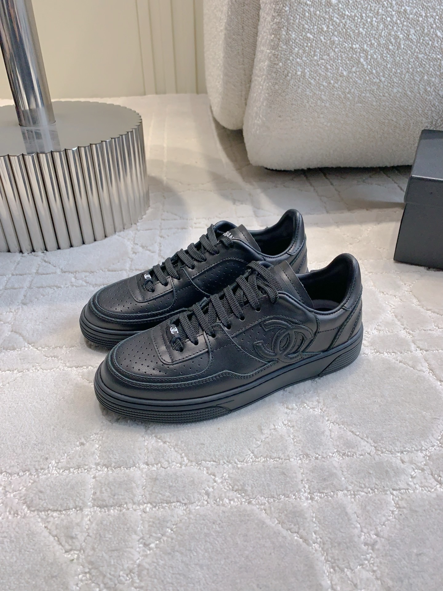 A pair of black Chanel leather sneakers featuring the interlocking CC logo and perforated details on a white textured carpet. - Hình 2