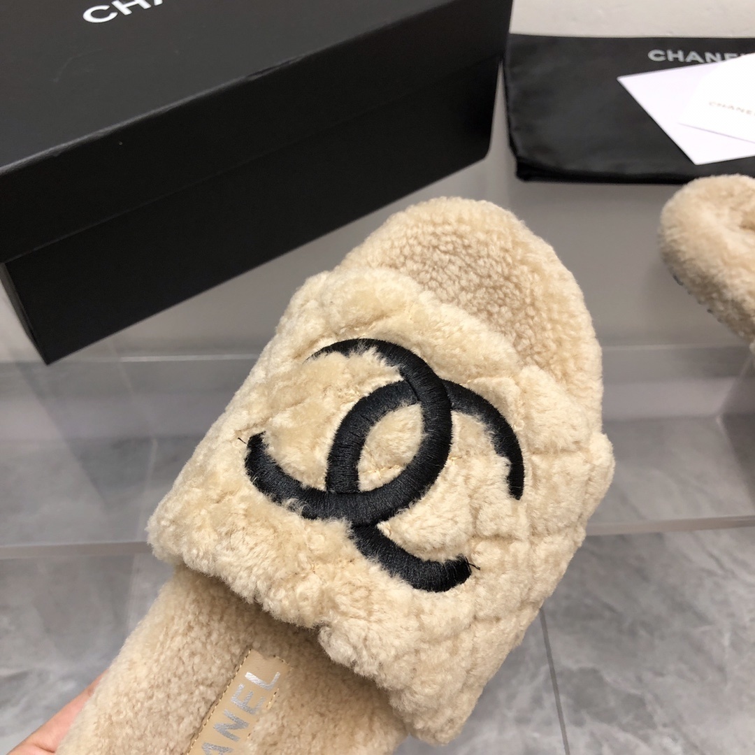 A pair of beige Chanel shearling quilted slides with a black CC logo displayed on a marble floor with its original box and dust bag. - Hình 4