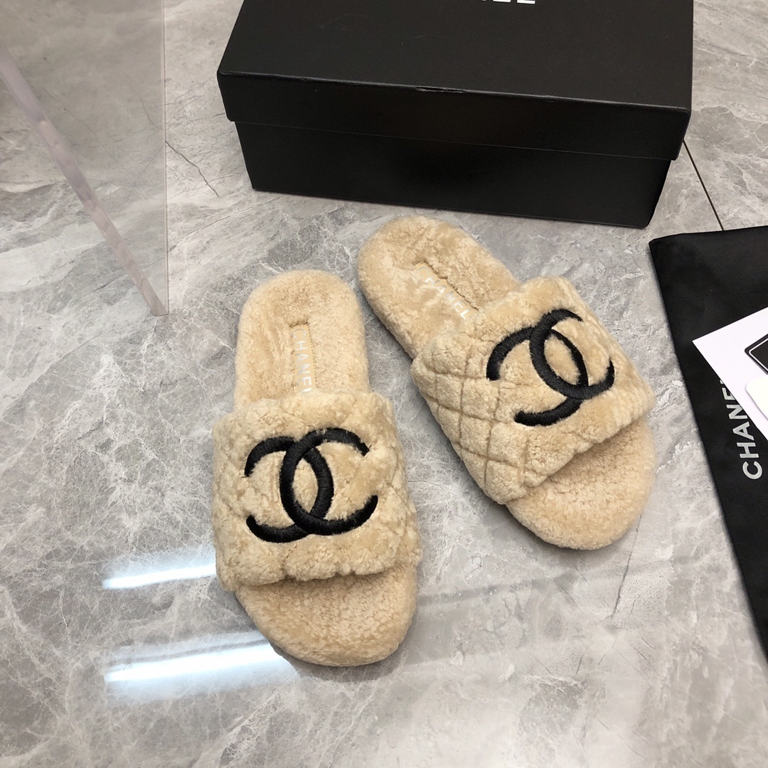 A pair of beige Chanel shearling quilted slides with a black CC logo displayed on a marble floor with its original box and dust bag. - Hình 3