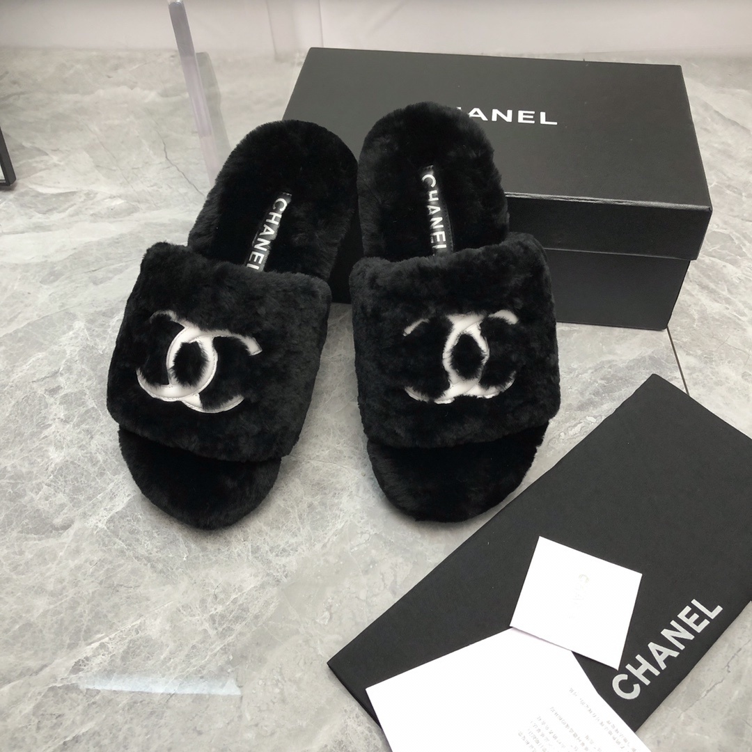 Pair of black fuzzy Chanel slippers with white interlocking CC logo on a glass table next to a designer box - Hình 9