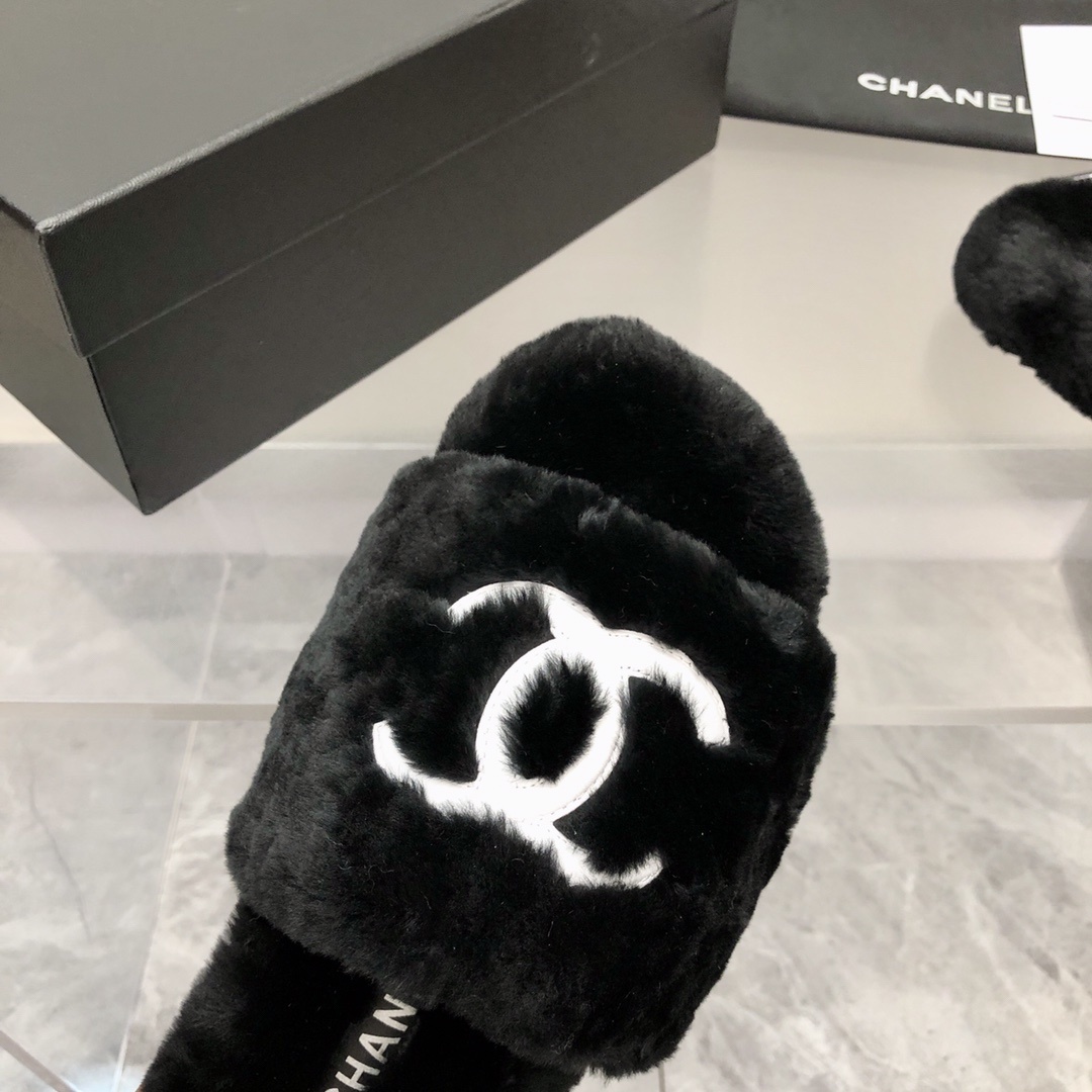 Pair of black fuzzy Chanel slippers with white interlocking CC logo on a glass table next to a designer box - Hình 5