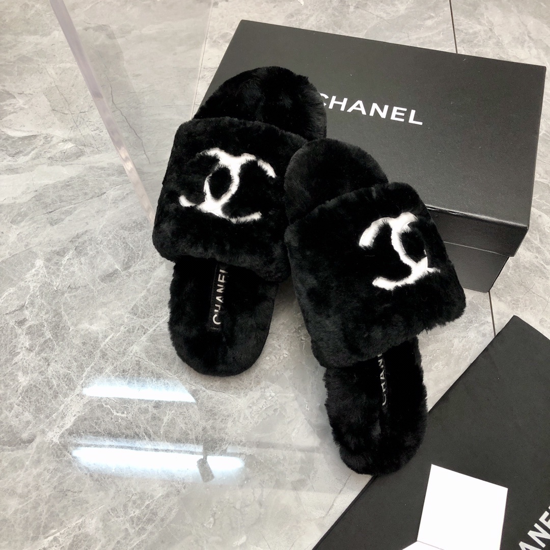 Pair of black fuzzy Chanel slippers with white interlocking CC logo on a glass table next to a designer box - Hình 4