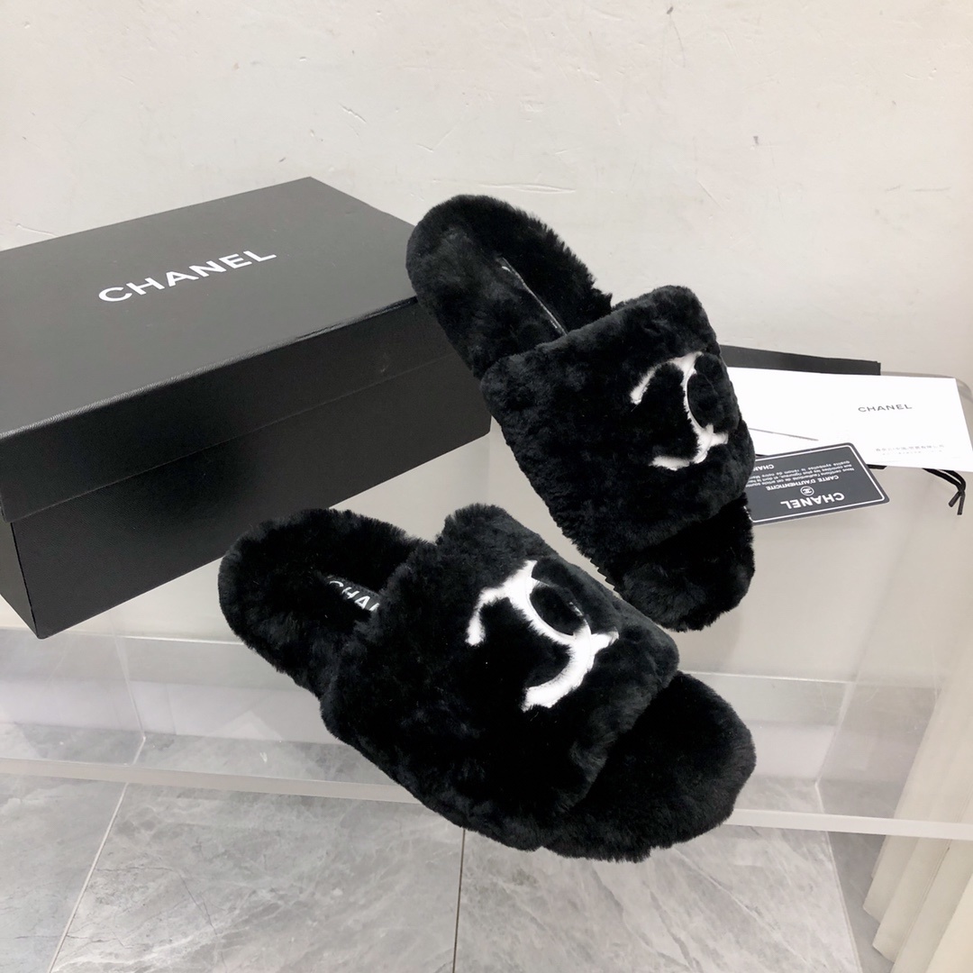 Pair of black fuzzy Chanel slippers with white interlocking CC logo on a glass table next to a designer box - Hình 3