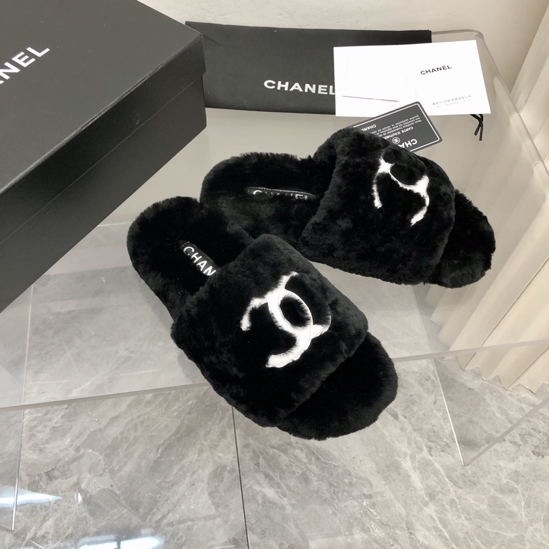 Pair of black fuzzy Chanel slippers with white interlocking CC logo on a glass table next to a designer box - Hình 2