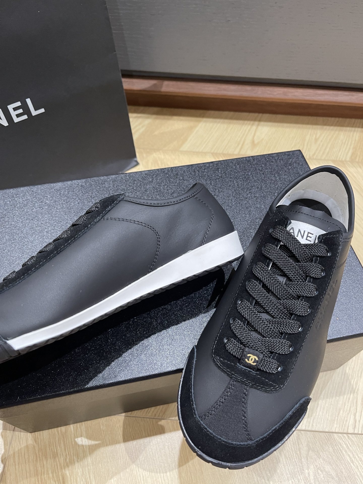 A pair of black Chanel low-top leather and suede sneakers with gold CC logos on the laces, sitting on a black Chanel shoe box. - Hình 8