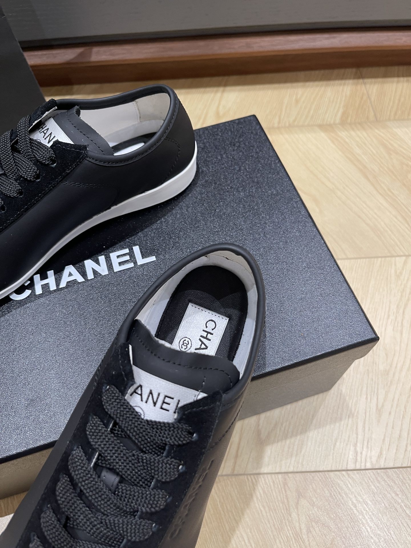 A pair of black Chanel low-top leather and suede sneakers with gold CC logos on the laces, sitting on a black Chanel shoe box. - Hình 7