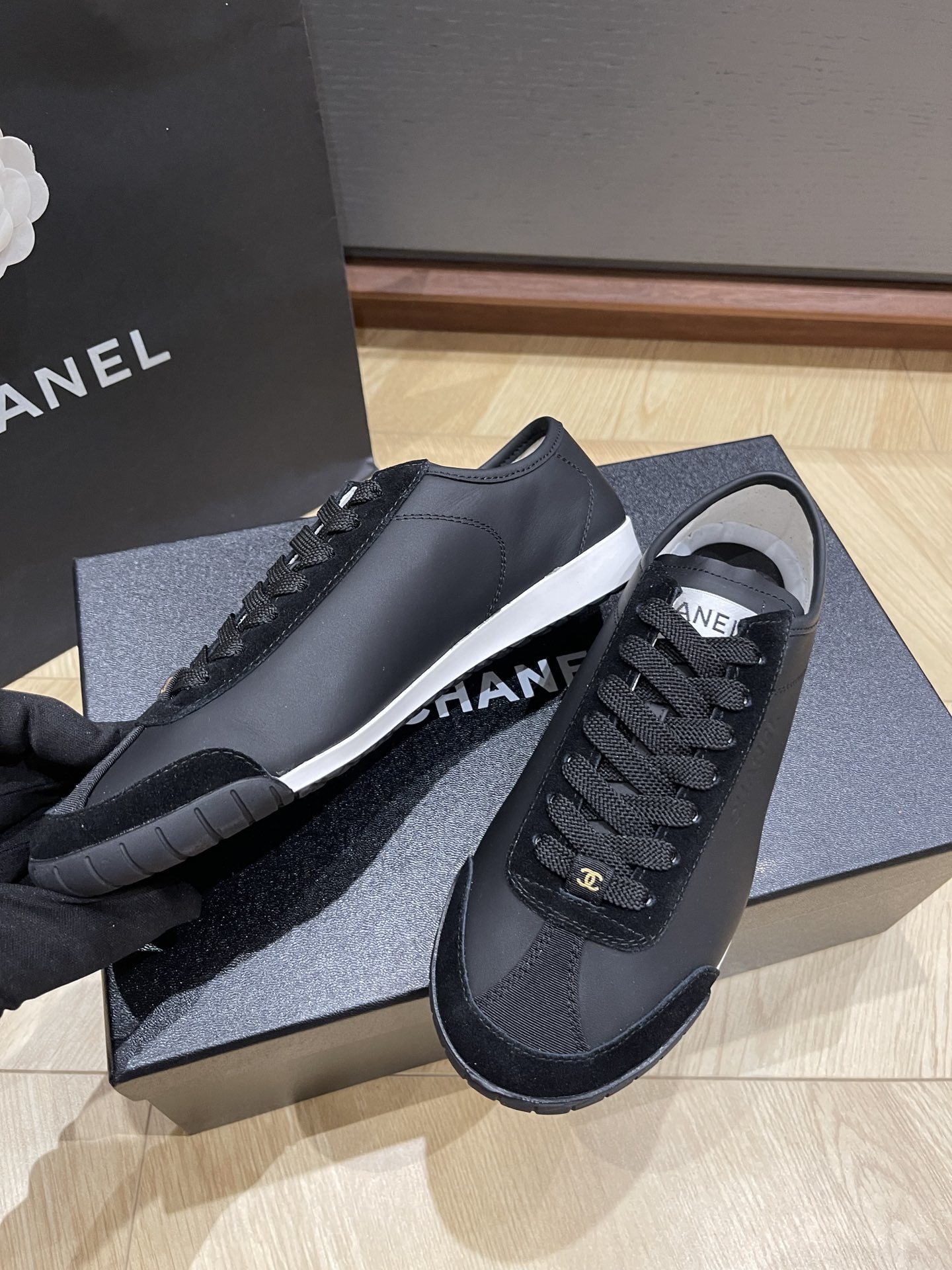 A pair of black Chanel low-top leather and suede sneakers with gold CC logos on the laces, sitting on a black Chanel shoe box. - Hình 6