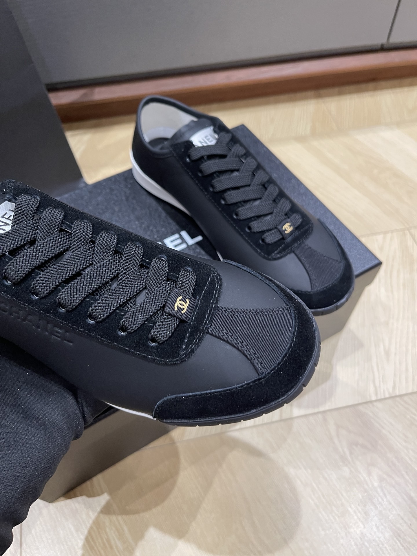 A pair of black Chanel low-top leather and suede sneakers with gold CC logos on the laces, sitting on a black Chanel shoe box. - Hình 5