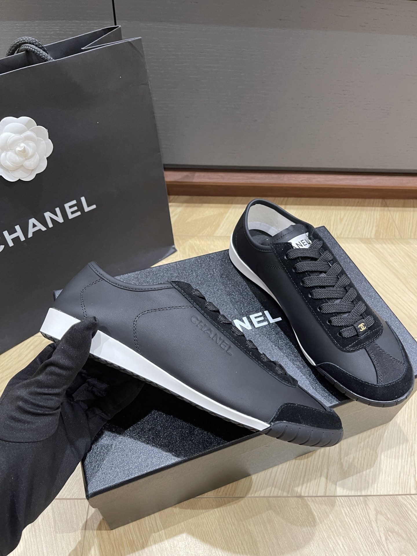 A pair of black Chanel low-top leather and suede sneakers with gold CC logos on the laces, sitting on a black Chanel shoe box. - Hình 4