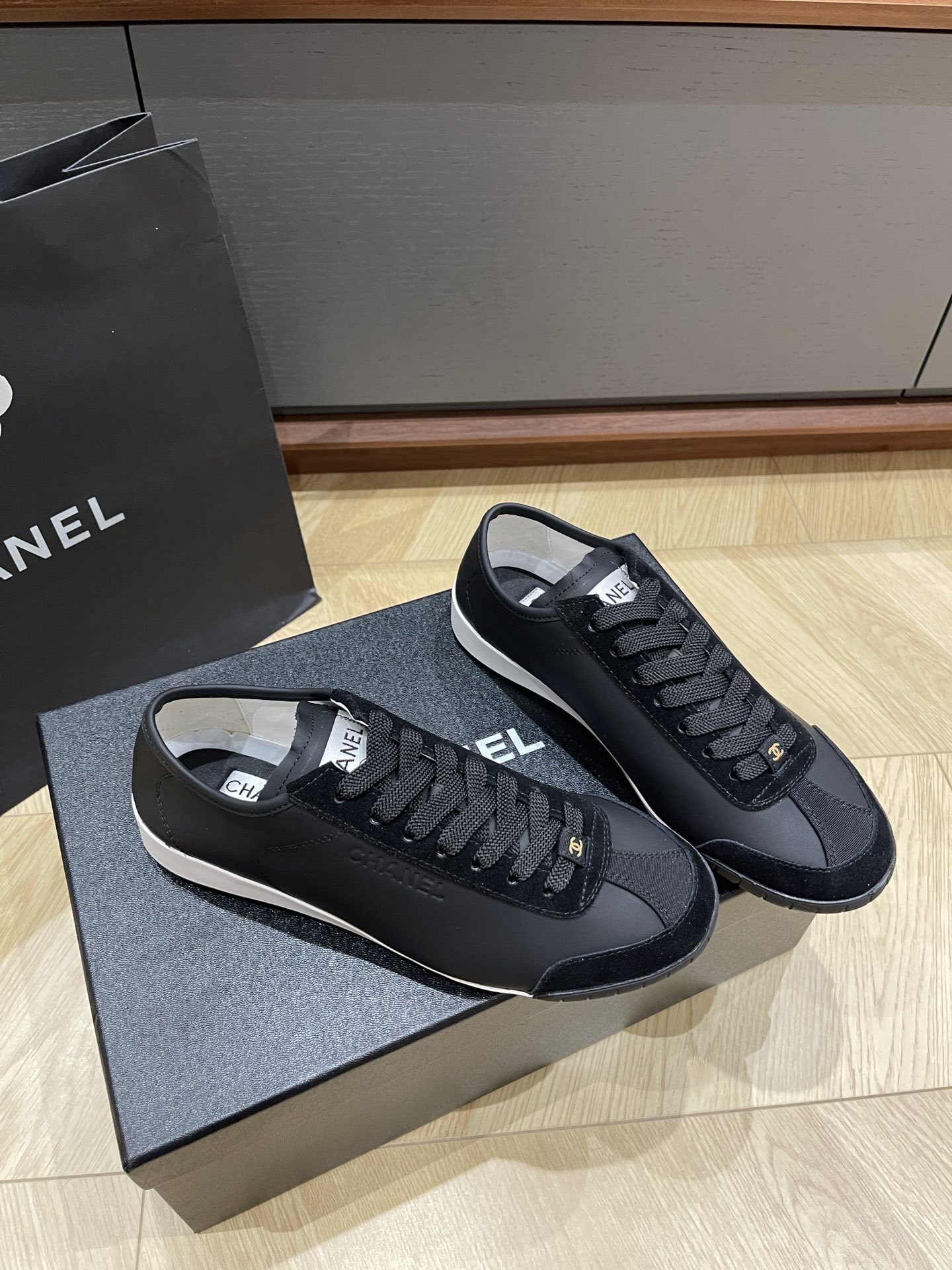 A pair of black Chanel low-top leather and suede sneakers with gold CC logos on the laces, sitting on a black Chanel shoe box. - Hình 3
