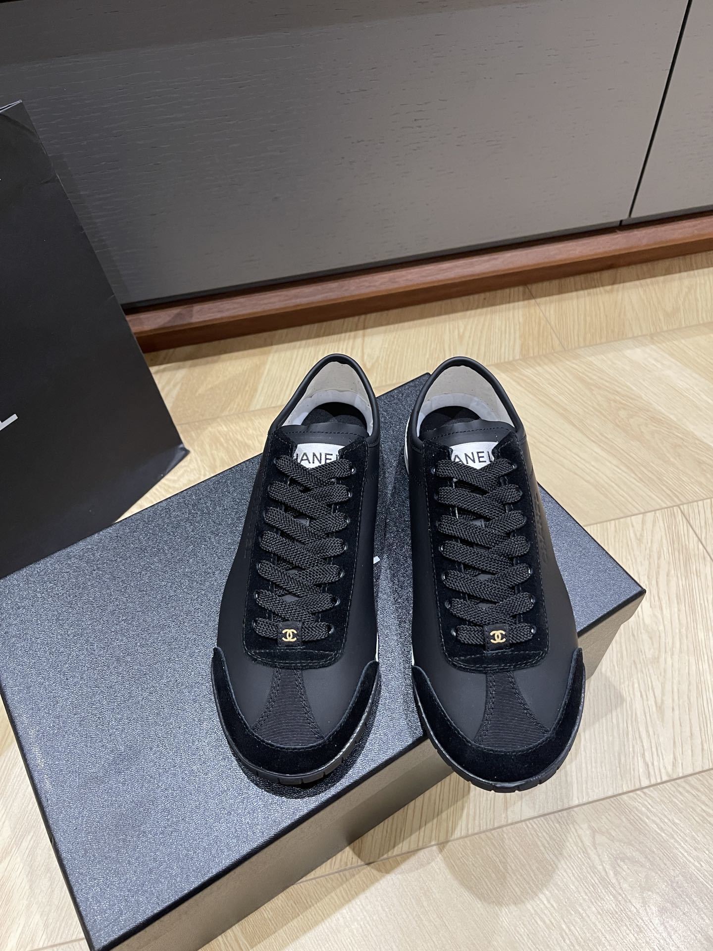 A pair of black Chanel low-top leather and suede sneakers with gold CC logos on the laces, sitting on a black Chanel shoe box. - Hình 2