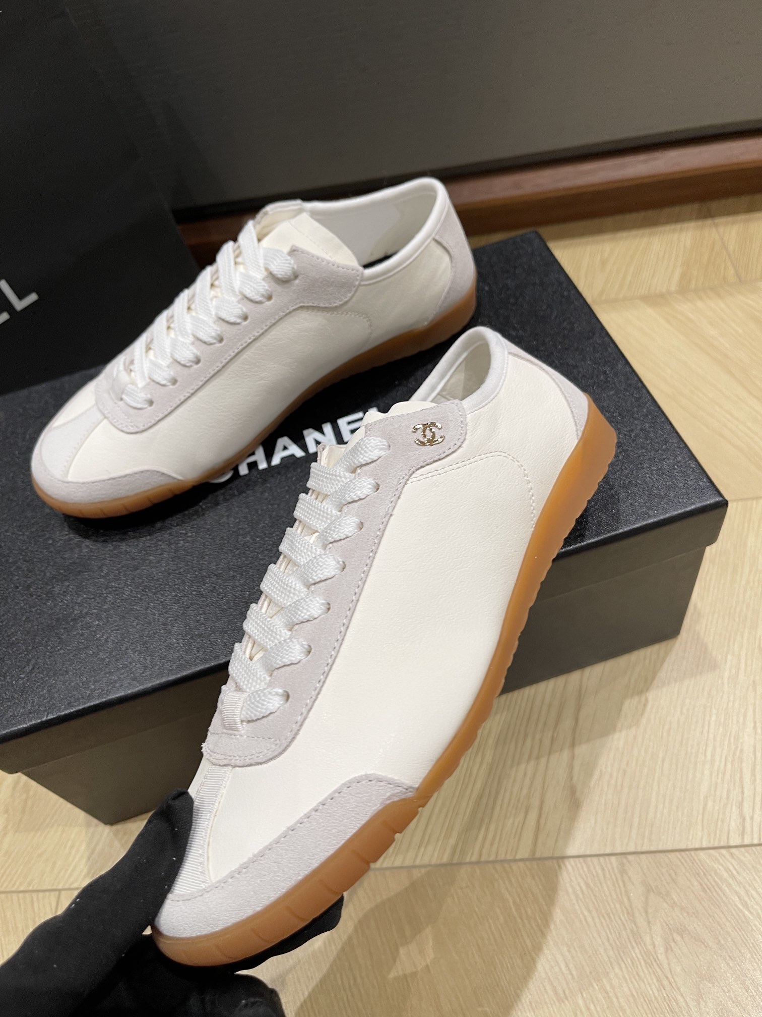 A pair of white Chanel low-top sneakers with suede trim and gum soles sitting on a black designer box next to a Chanel shopping bag. - Hình 7