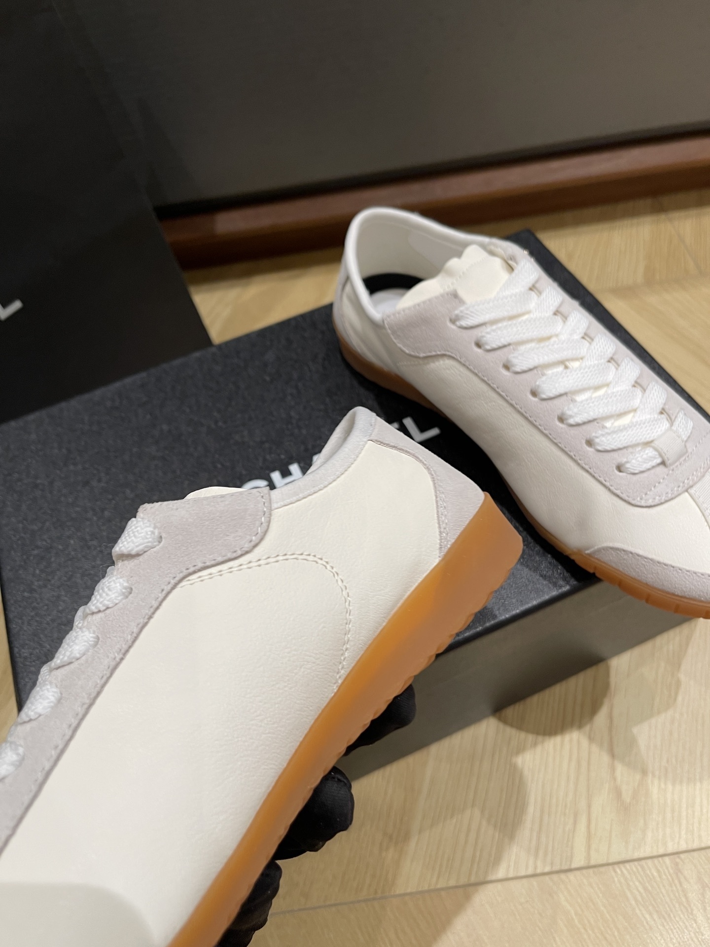 A pair of white Chanel low-top sneakers with suede trim and gum soles sitting on a black designer box next to a Chanel shopping bag. - Hình 6