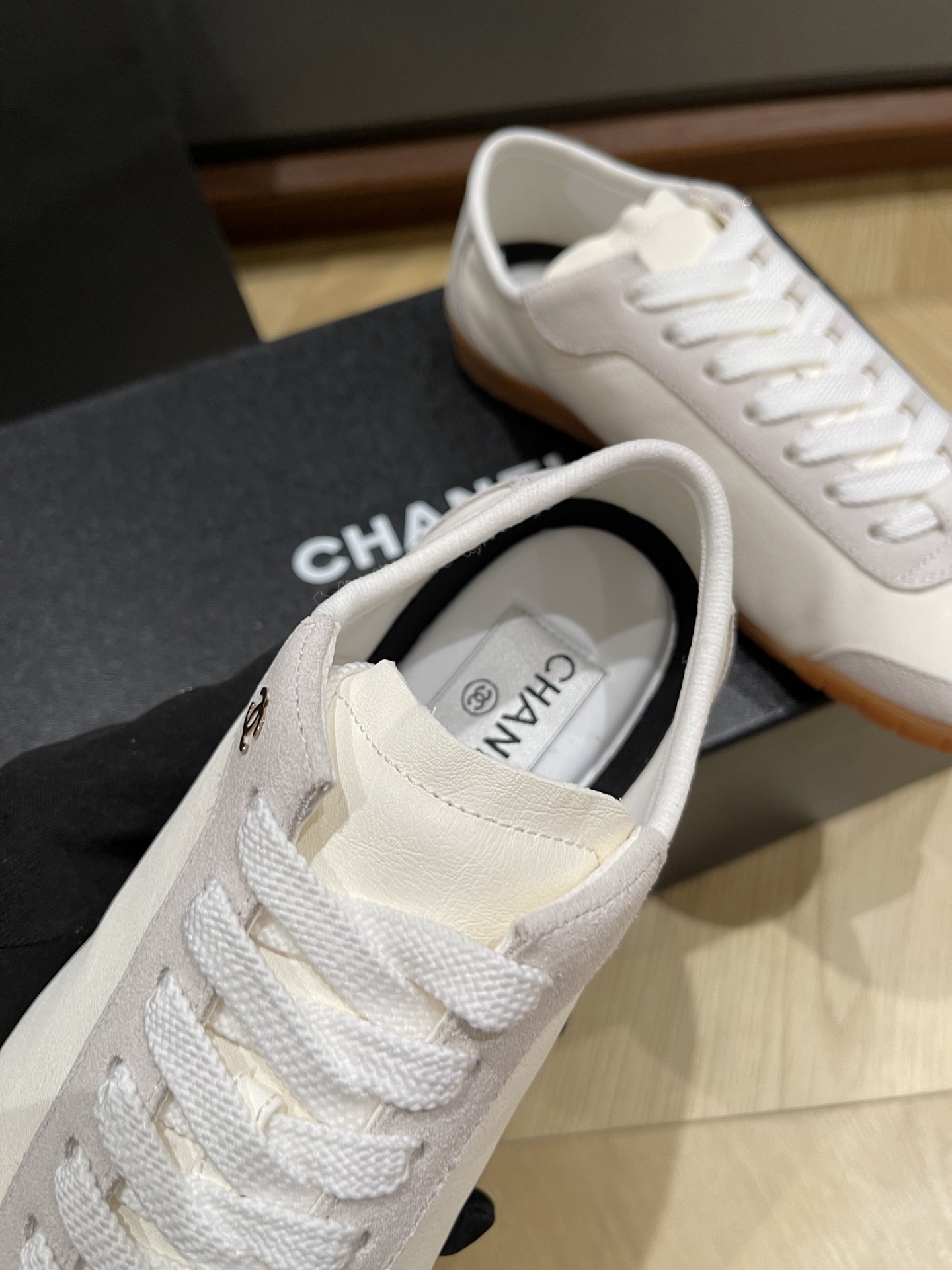 A pair of white Chanel low-top sneakers with suede trim and gum soles sitting on a black designer box next to a Chanel shopping bag. - Hình 5