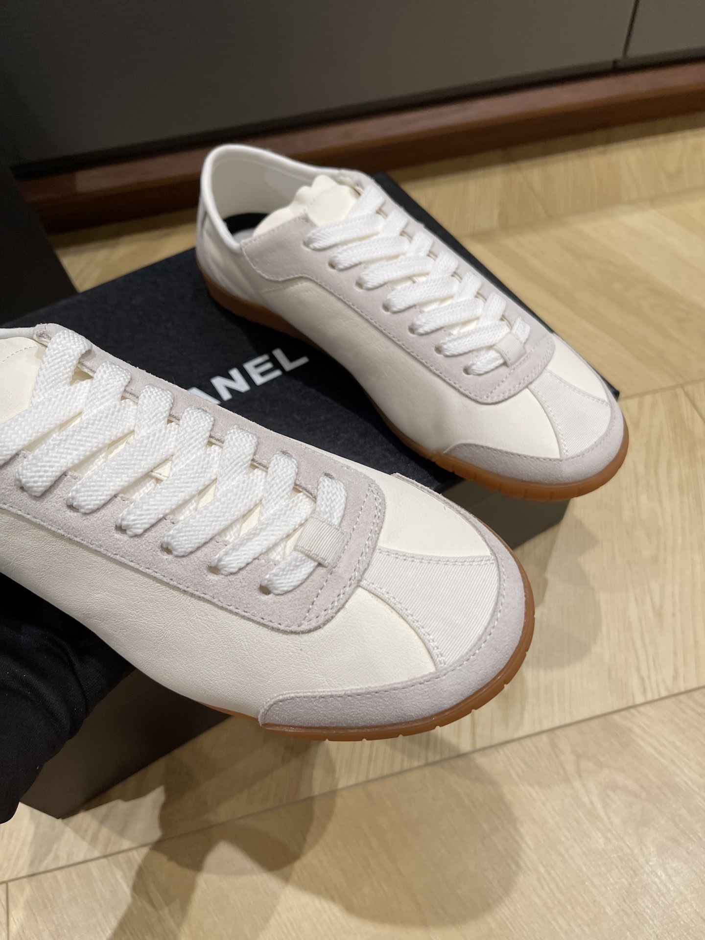 A pair of white Chanel low-top sneakers with suede trim and gum soles sitting on a black designer box next to a Chanel shopping bag. - Hình 4