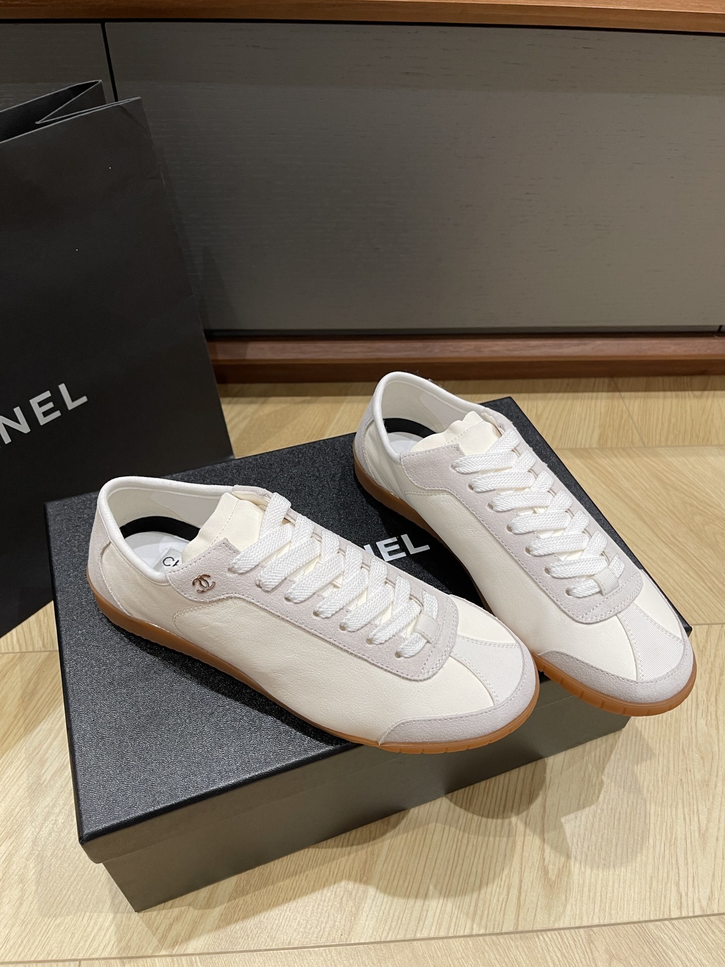 A pair of white Chanel low-top sneakers with suede trim and gum soles sitting on a black designer box next to a Chanel shopping bag. - Hình 2