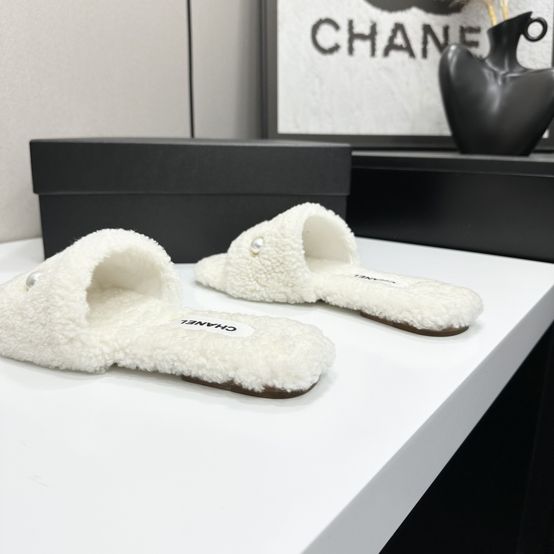 A pair of white Chanel shearling slippers featuring pearl and gold CC logo embellishments sitting on a black designer box. - Hình 9