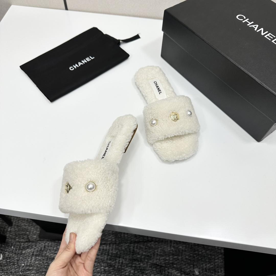 A pair of white Chanel shearling slippers featuring pearl and gold CC logo embellishments sitting on a black designer box. - Hình 7