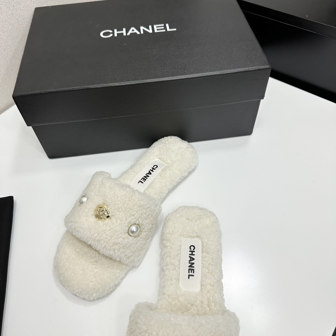 A pair of white Chanel shearling slippers featuring pearl and gold CC logo embellishments sitting on a black designer box. - Hình 6